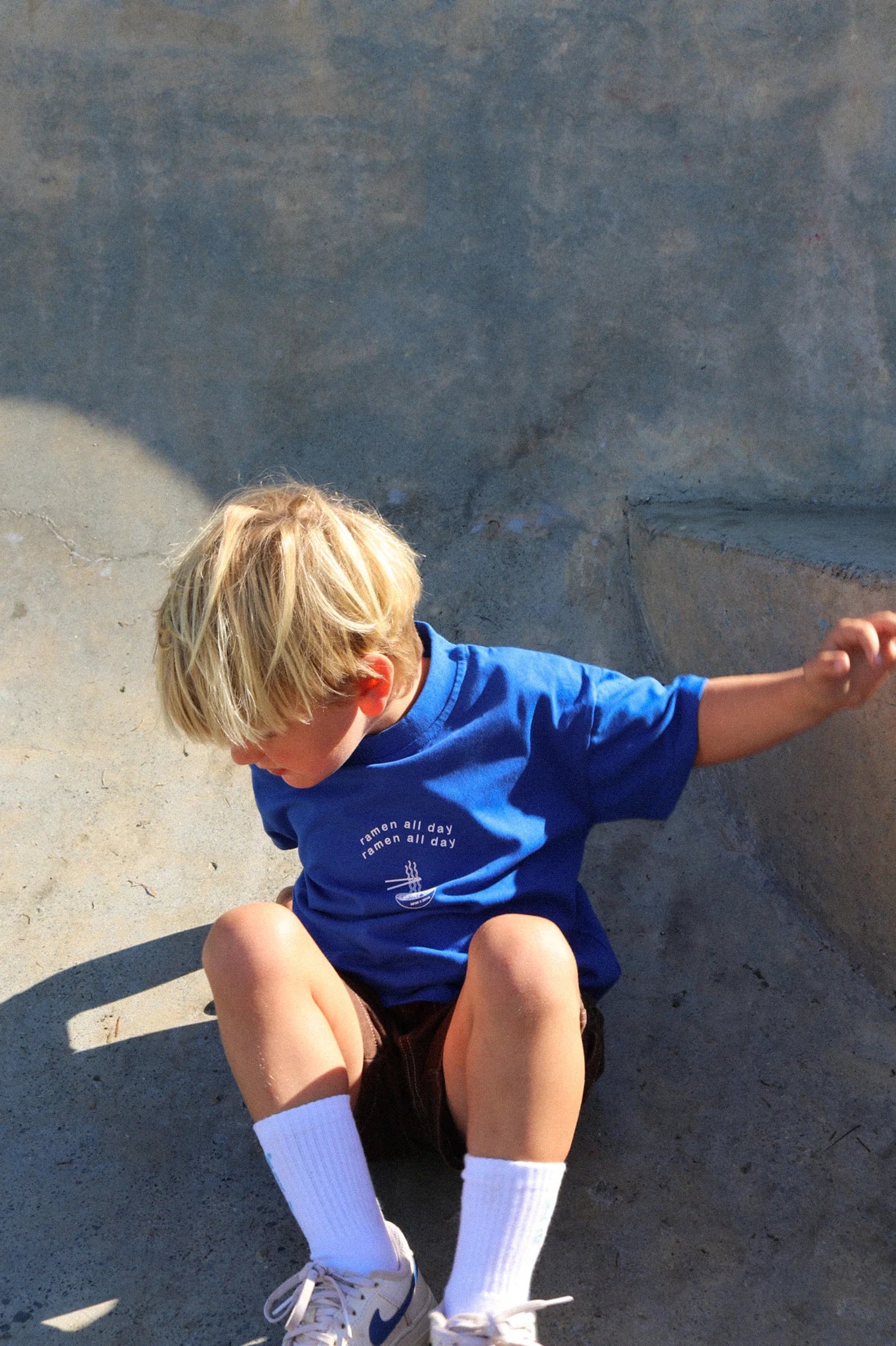 A young blond child sits against a concrete wall wearing a ZEPHII Ramen All Day Tee, dark shorts, white socks, and sneakers. Looking down with one hand on the wall, they bask in the sunny outdoor vibe.