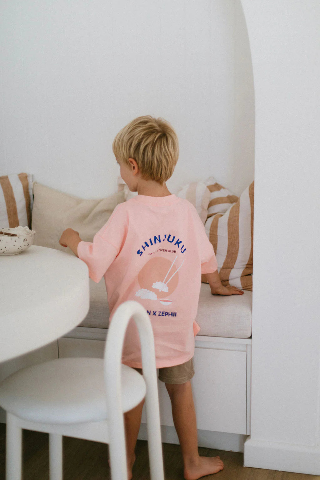 A blonde child in an oversized ZEPHII Shinjuku Tee stands in a cozy, light-colored room with striped bench cushions, facing away and gazing at the wall, embodying vibrant Japanese culture.