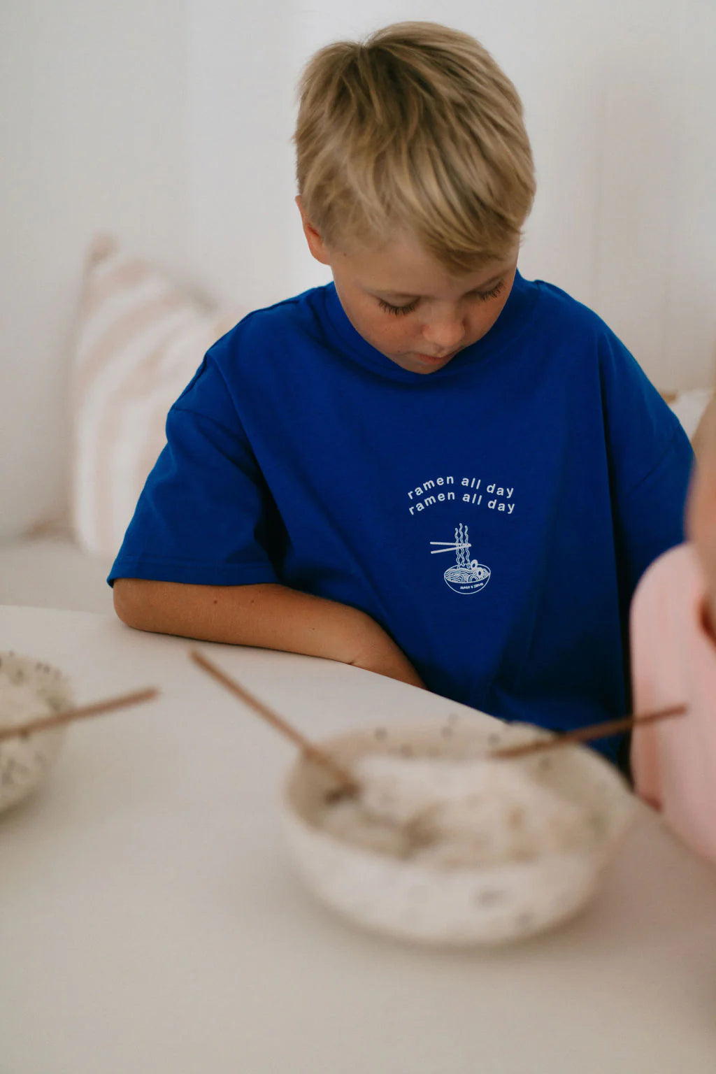 A blond-haired child is seated at a table, wearing ZEPHII's vintage cool "Ramen All Day Tee." Two bowls with chopsticks on the table reflect a laid-back nod to Japanese culture.