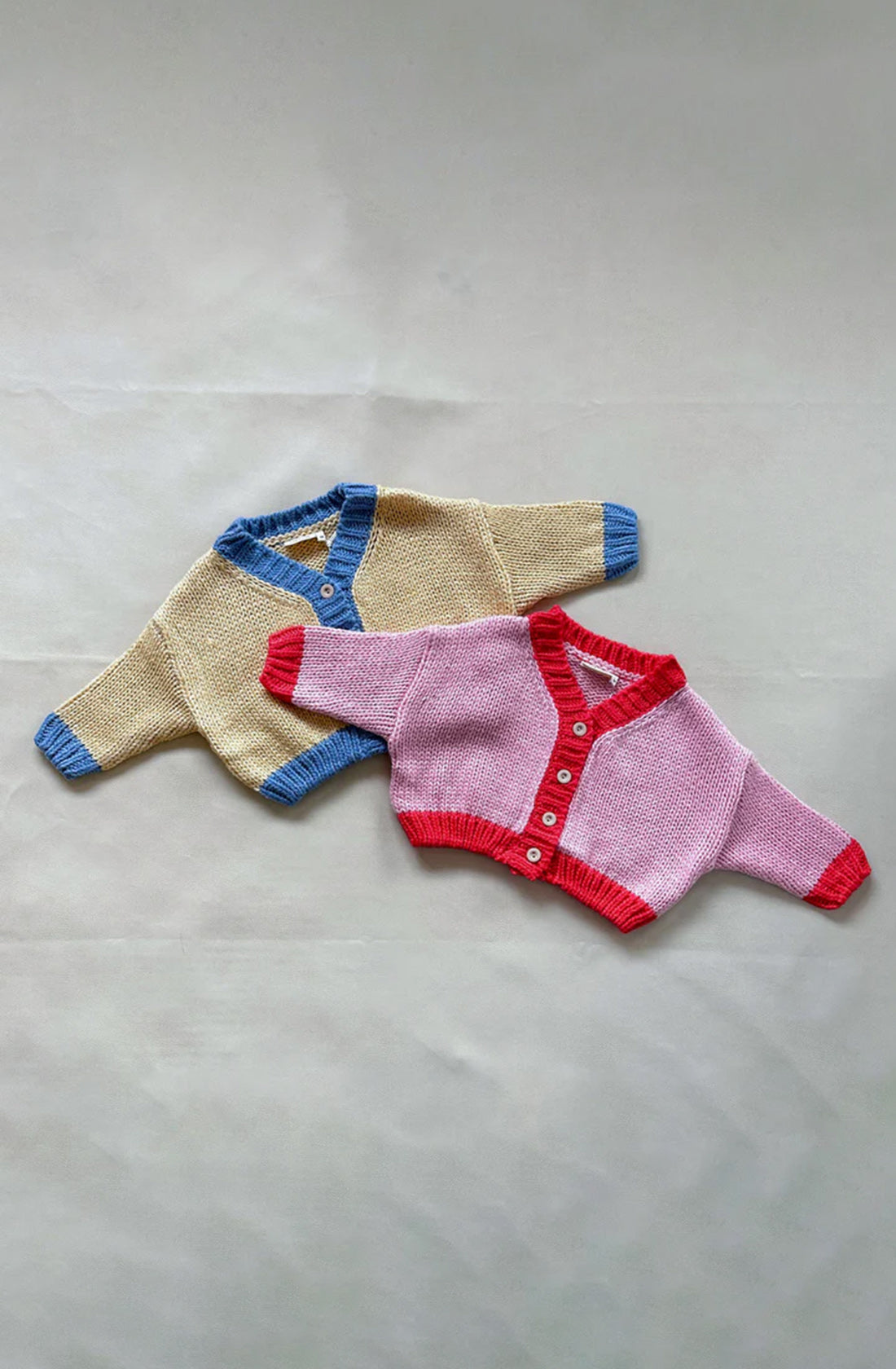 Two hand-knitted baby cardigans from TINY TROVE are lying flat on a grey surface. The top cardigan, with an oversized fit, is cream-colored with blue trim. The bottom Zuri Contrast Knit Cardigan in light pink with red trim showcases the cozy craftsmanship of TINY TROVE. Both chunky knit cardigans feature long sleeves and buttons down the front.