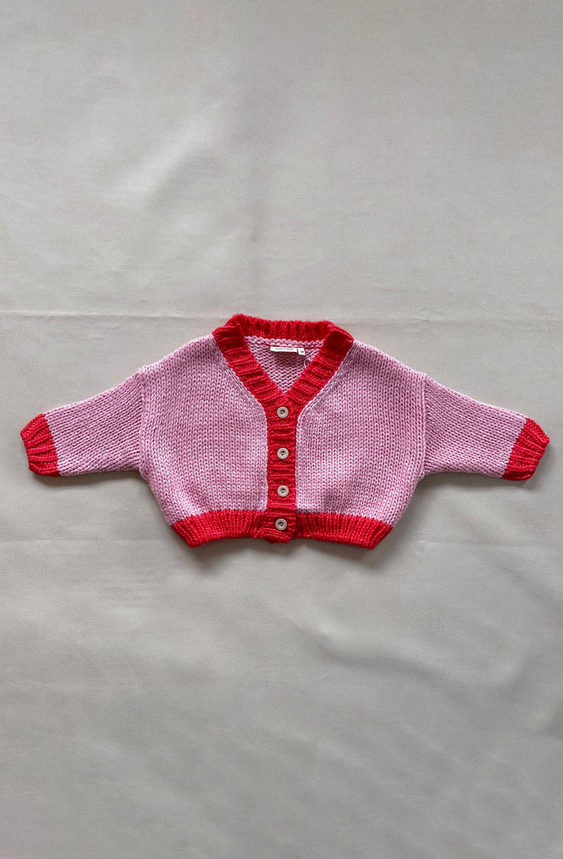 A small Zuri Contrast Knit Cardigan Pink/Red by TINY TROVE with long sleeves lies flat. The chunky knit cardigan features a primarily light pink color with vibrant red accents on the edges of the sleeves, collar, and buttoned front placket. It has five buttons down the front and is made from recycled acrylic.