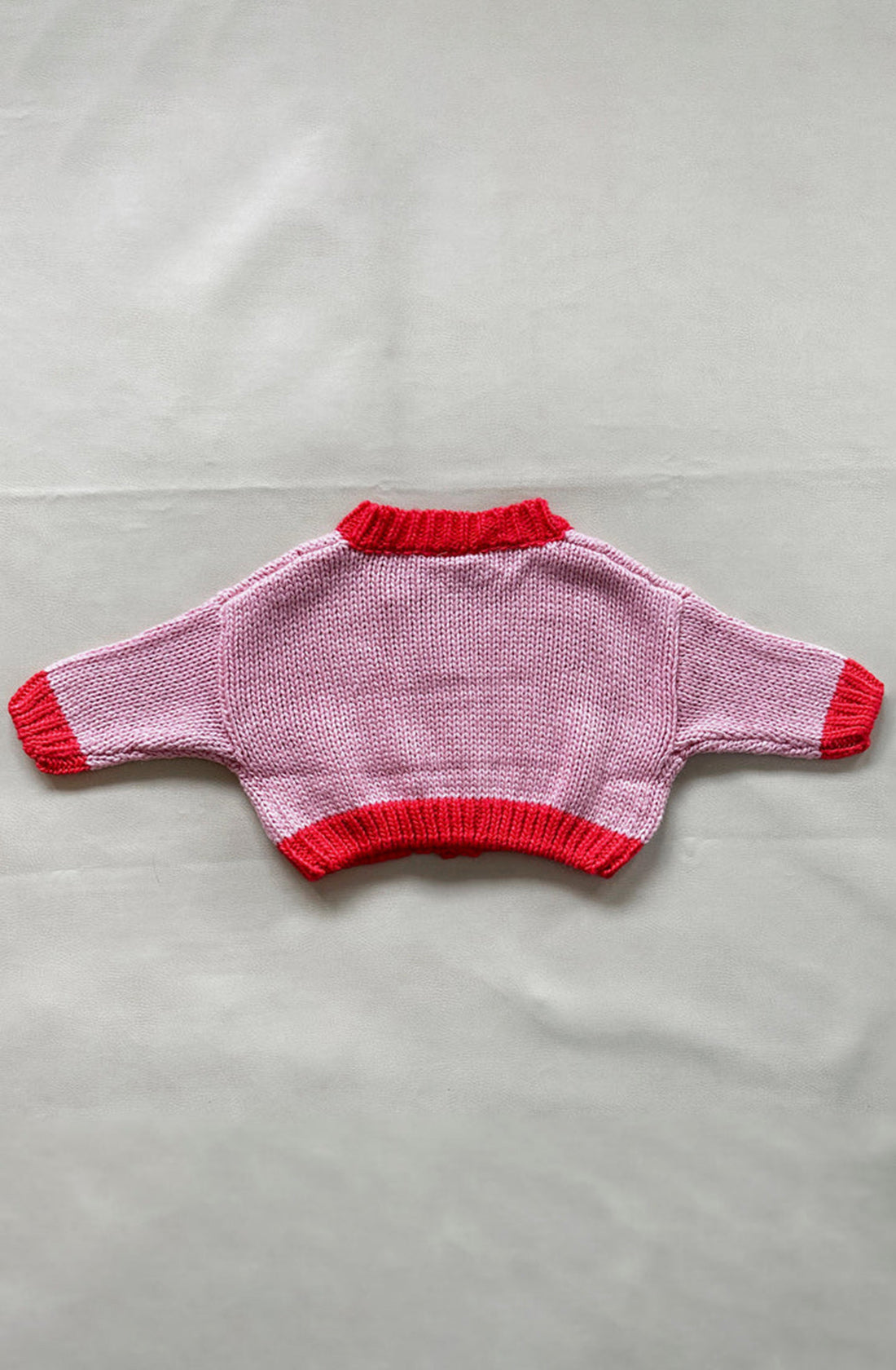 Introducing the Zuri Contrast Knit Cardigan in Pink/Red by TINY TROVE, featuring a charming pink and red checkered pattern. This hand-knitted baby sweater is crafted from recycled acrylic, with solid red detailing on the collar, cuffs, and hem. The chunky knit design adds texture and warmth. Laid flat on a neutral background, its oversized fit beautifully showcases its full design and long sleeves.