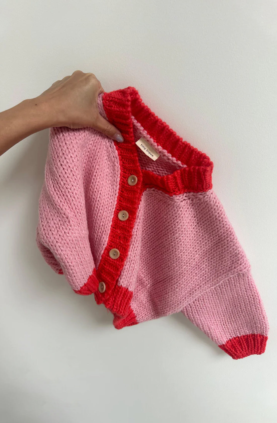 A hand is holding the Zuri Contrast Knit Cardigan in Pink/Red by TINY TROVE, showcasing its chunky knit design with red trim and wooden buttons against a white background. This eco-friendly cardigan is crafted from recycled acrylic and features a ribbed collar and hem.