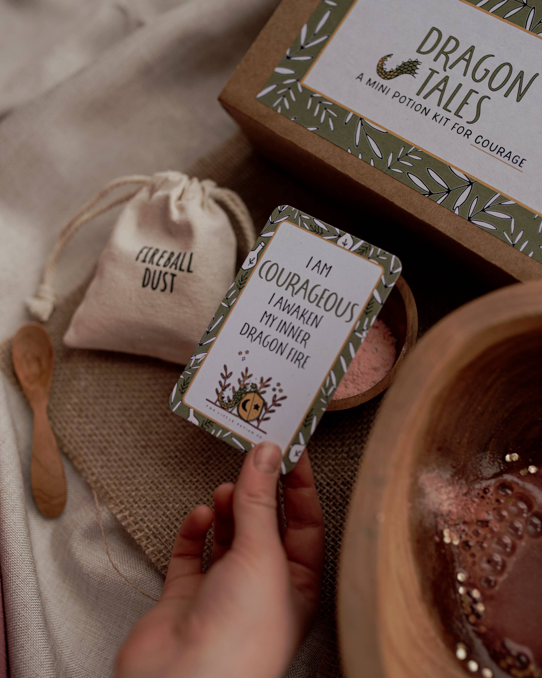 A hand holds a card that reads, "I am courageous, I awaken my inner dragon fire!" Next to it is a small drawstring bag labeled "Fireball dust" and a wooden bowl filled with pink powder. All of this is set on a burlap surface alongside a box titled "MINI Dragon Tales Potion Kit (A potion for courage)" by THE LITTLE POTION CO. This imaginative play potion kit transports you to the world of dragons and knights.