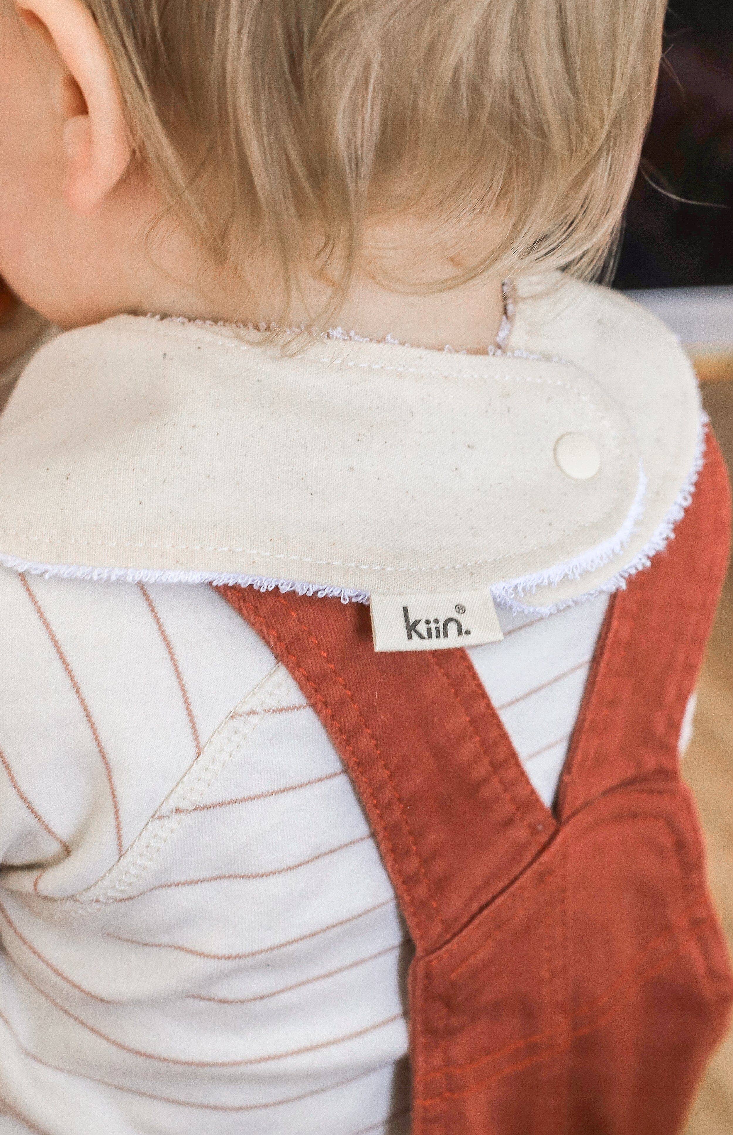 A blond child wears a cream cotton "Dribble Bib Oat" by KIIN BABY and a red-brown overall. A tag on the bib reads "kiin." Snap fasteners keep it secure as the child slightly turns, showing a close-up of their head and upper body.