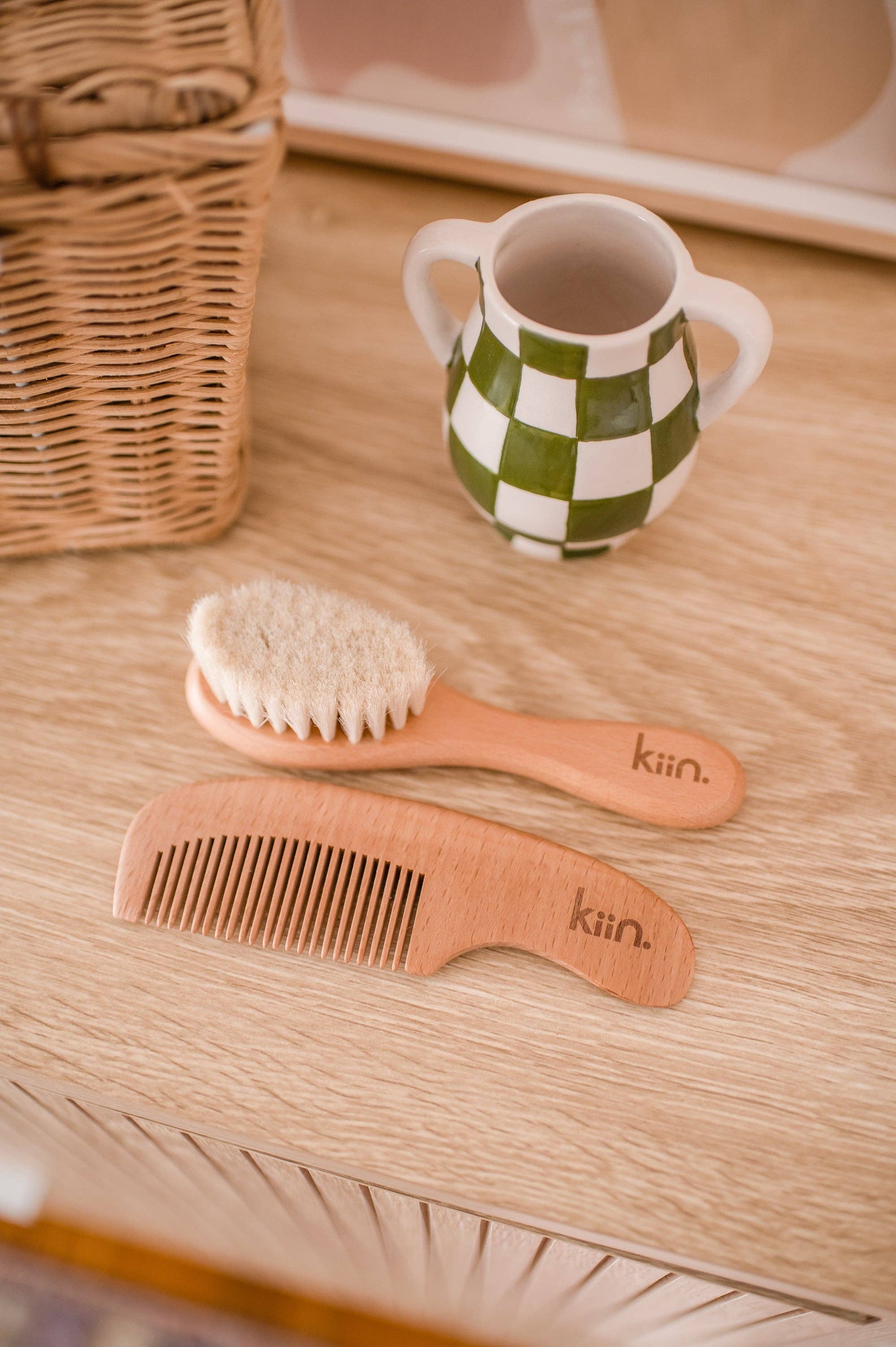 A Wooden Baby Brush and Comb Set by KIIN BABY lies on a light wood surface, alongside an eco-friendly ceramic cup featuring a green and white checkered pattern. A wicker basket peeks into the scene.