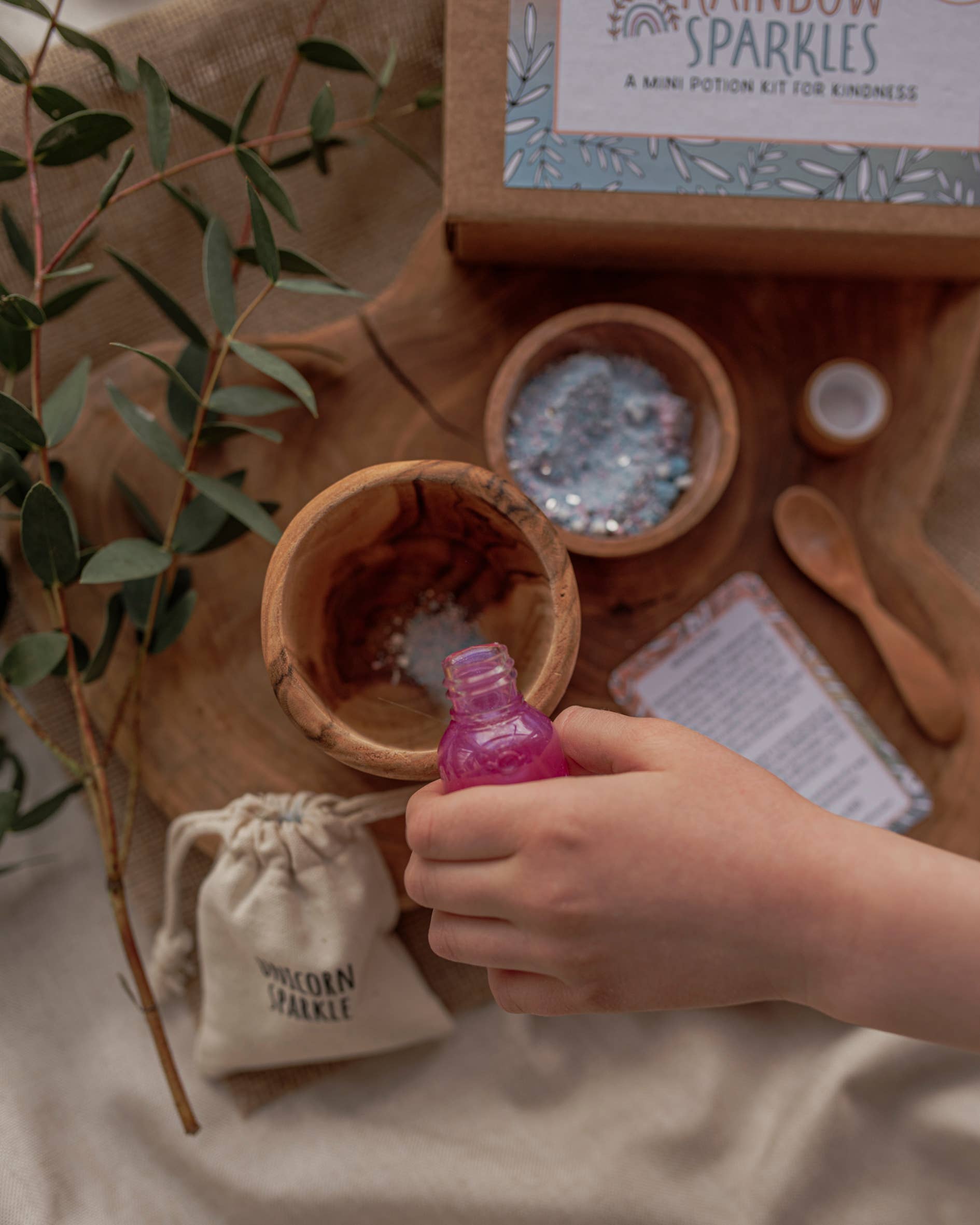 A child's hand holds a small pink bottle over a wooden bowl containing sparkly materials. Nearby are a wooden dish with blue glitter, a recipe card, and a cloth bag labeled "UNICORN SPARKLES." In the background, there's a box labeled "MINI Rainbow Sparkles Potion Kit (A potion for kindness)" by THE LITTLE POTION CO.