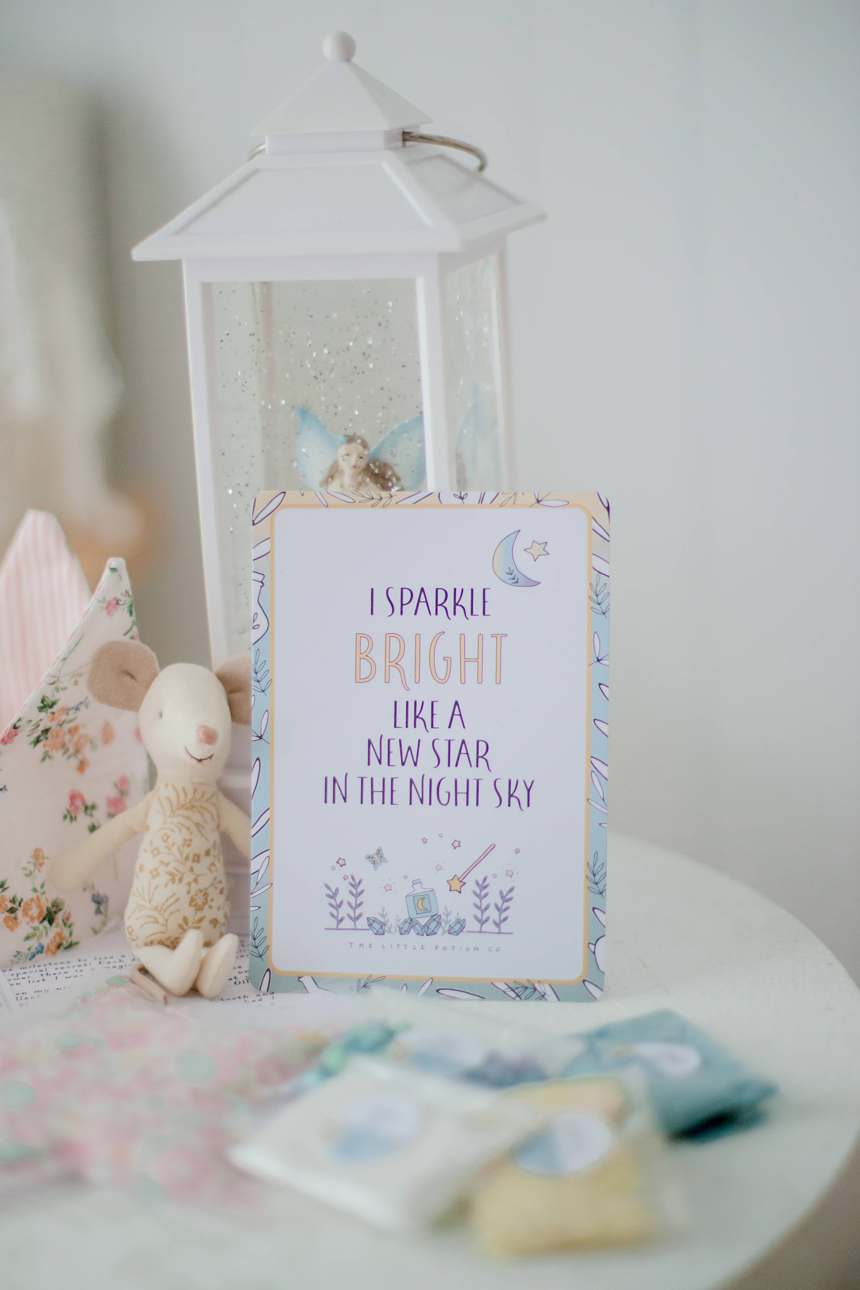 An affirmation card with the message "I sparkle bright like a new star in the night sky" is arranged on a round table. Surrounding it, a Tooth Fairy Magic Potion Pouch by THE LITTLE POTION CO, along with a white lantern, plush toy, and floral fabric pieces, creates a cozy and whimsical atmosphere evoking the lore of Tooth Fairy Magic Potion.