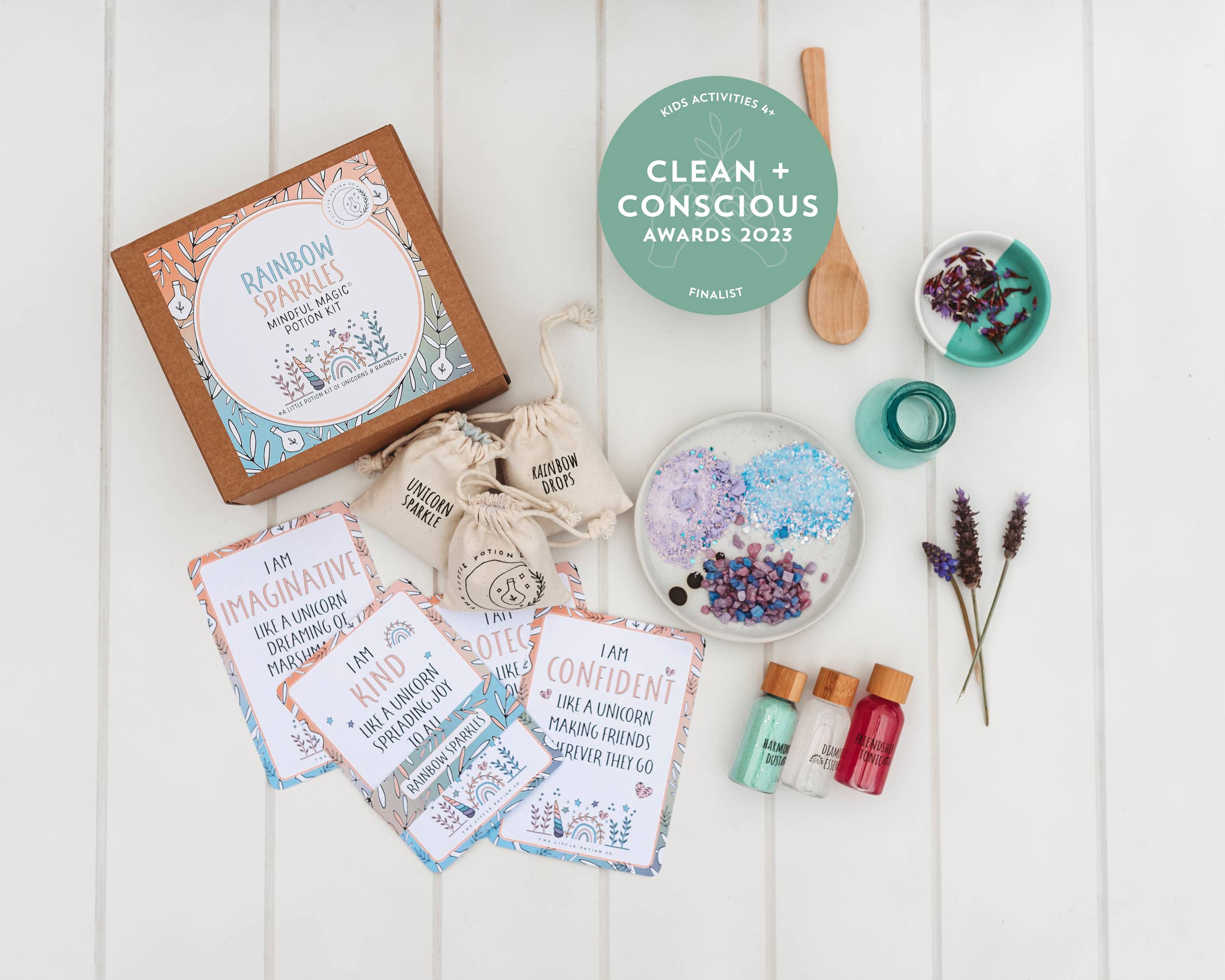 A flat lay showcasing the Rainbow Sparkles MINDFUL Potion Kit by THE LITTLE POTION CO, featuring affirmation cards, bottles of colored liquids, a wooden spoon, sachets labeled with words, and dried flowers. This enchanting kit also includes a circular badge for finalists in the Clean + Conscious Awards 2023.