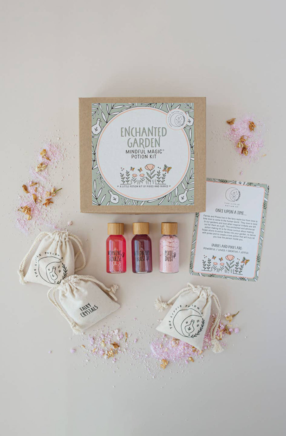 The image showcases an "Enchanted Garden MINDFUL Potion Kit" from THE LITTLE POTION CO. This potion-making kit includes three bottles of flower elixirs, a set of small drawstring pouches, and a detailed instruction card, all neatly arranged on a light background scattered with colorful powder.