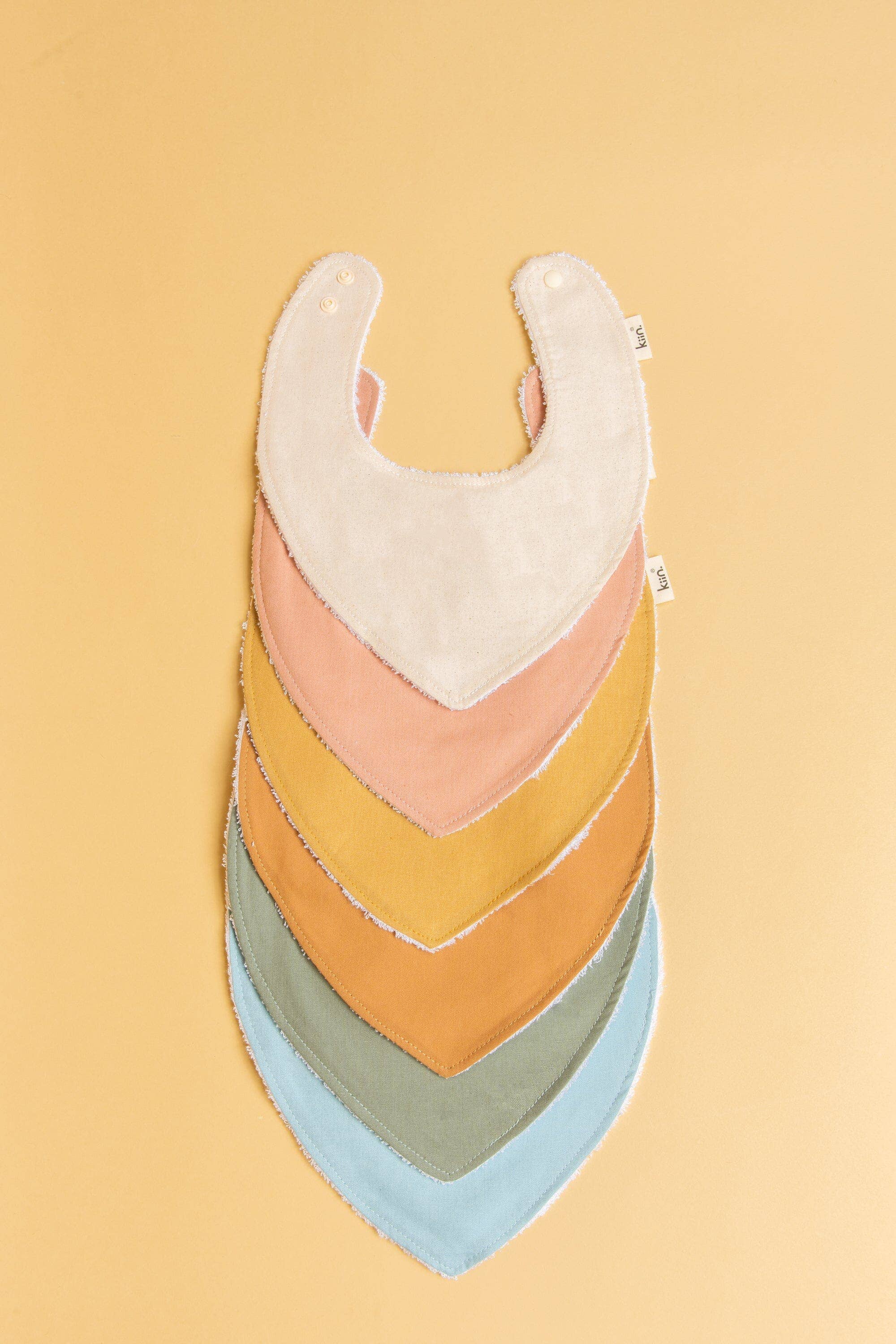 A stack of six KIIN BABY "Dribble Bib Oat" cotton bibs with snap fasteners, arranged in a fan shape on a beige background. They are layered from top to bottom in cream, pink, mustard, brown, green, and light blue hues.