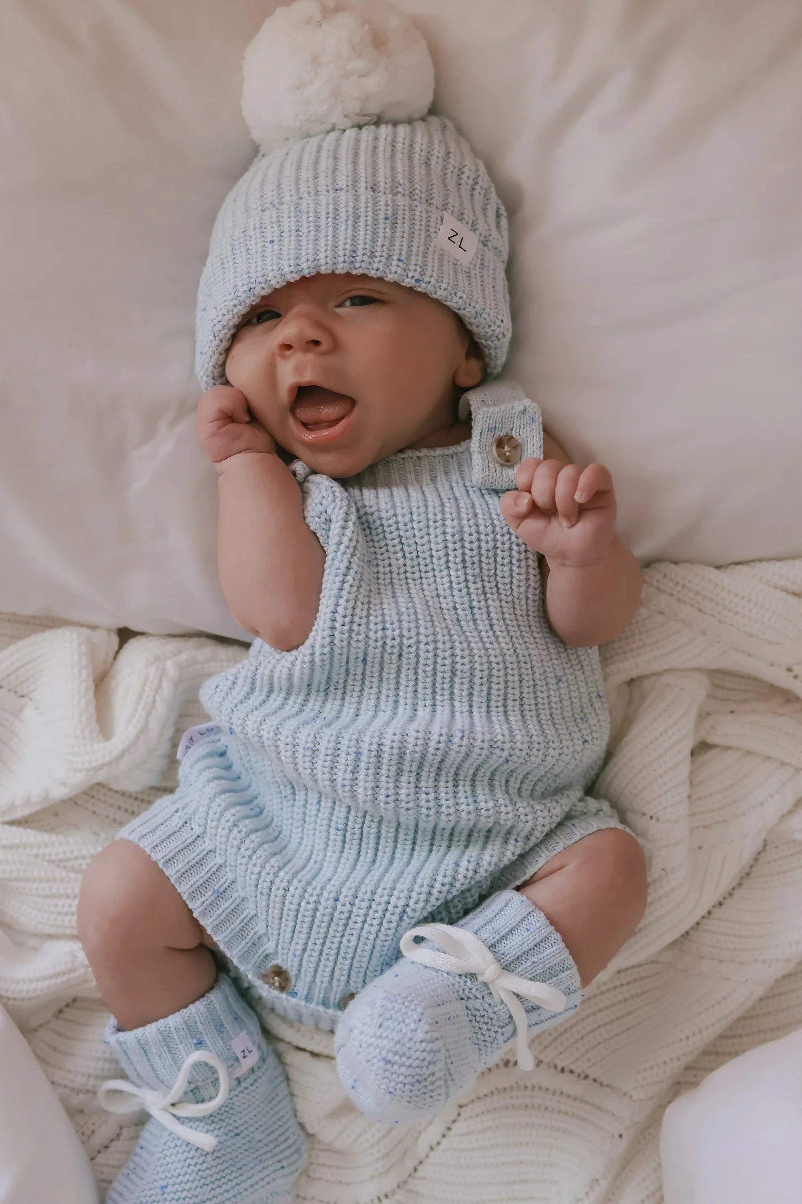 A baby wearing a light blue ZIGGY LOU Aero Fleck knitted outfit lies on a white blanket. The ensemble includes a hat with a pom-pom, sleeveless romper, and matching Booties Aero Fleck. With one hand near their face and the other holding a small square object, the baby looks absolutely adorable.