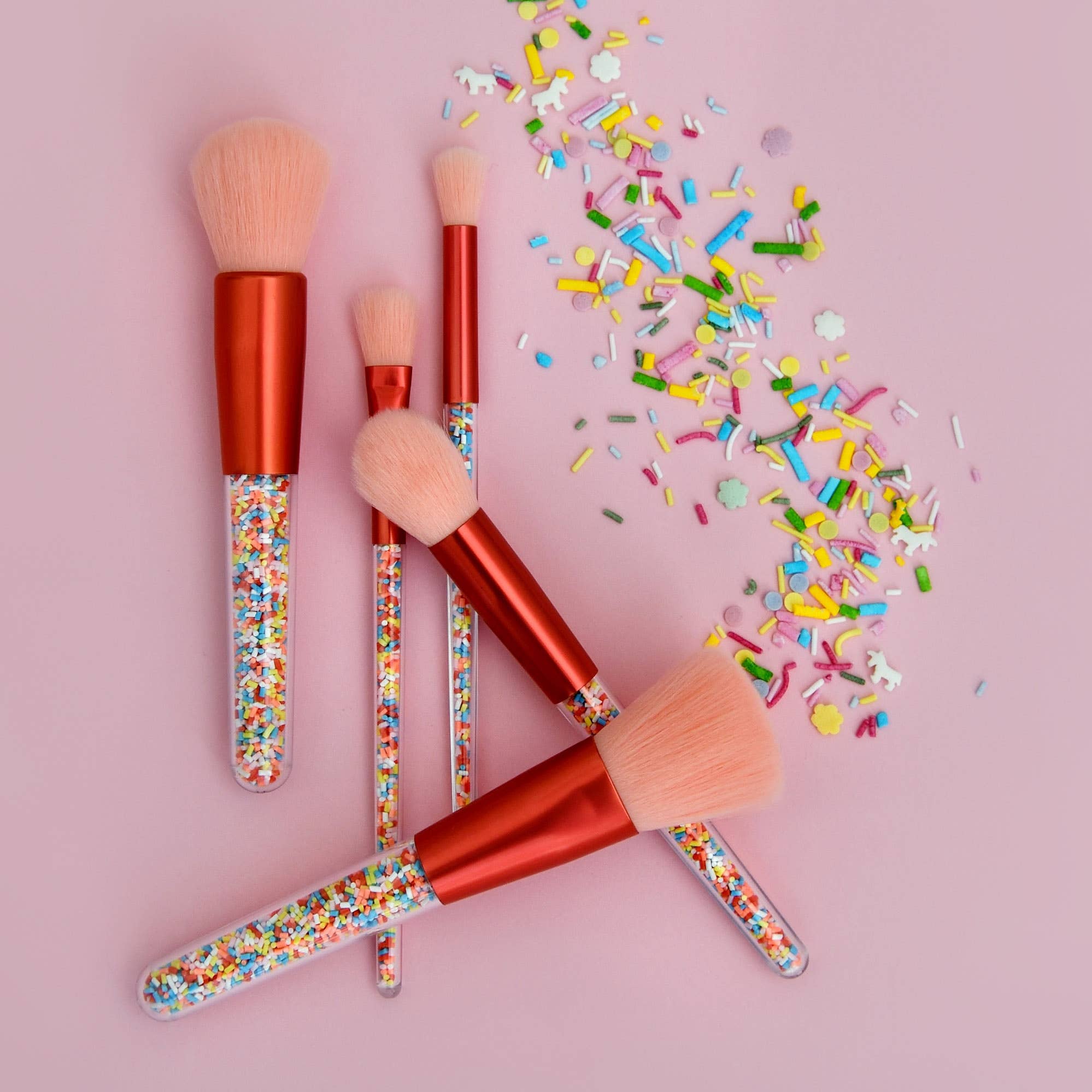 The Twinkle Sprinkle Brush Set by no nasties kids features hypoallergenic makeup brushes with light pink bristles and red handles adorned with colorful sprinkles on the lower part. The set is arranged on a pink background, with loose, rainbow-colored sprinkles scattered beside them.