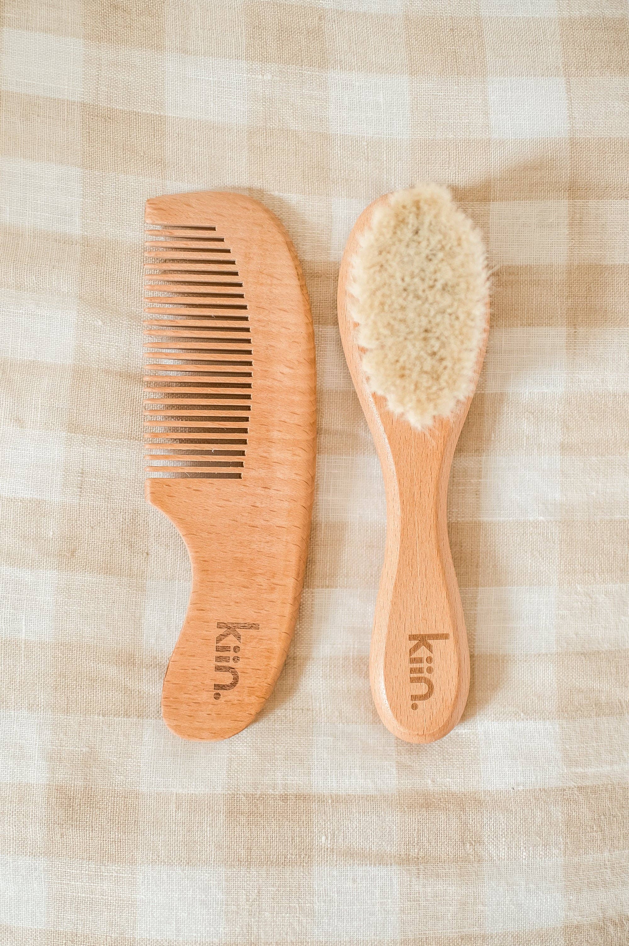 A Wooden Baby Brush and Comb Set from KIIN BABY rests on beige and white checkered fabric. Both feature "kin." engraved on the handles. The brush is crafted with soft, light-colored goat wool bristles for gentle care of delicate strands.