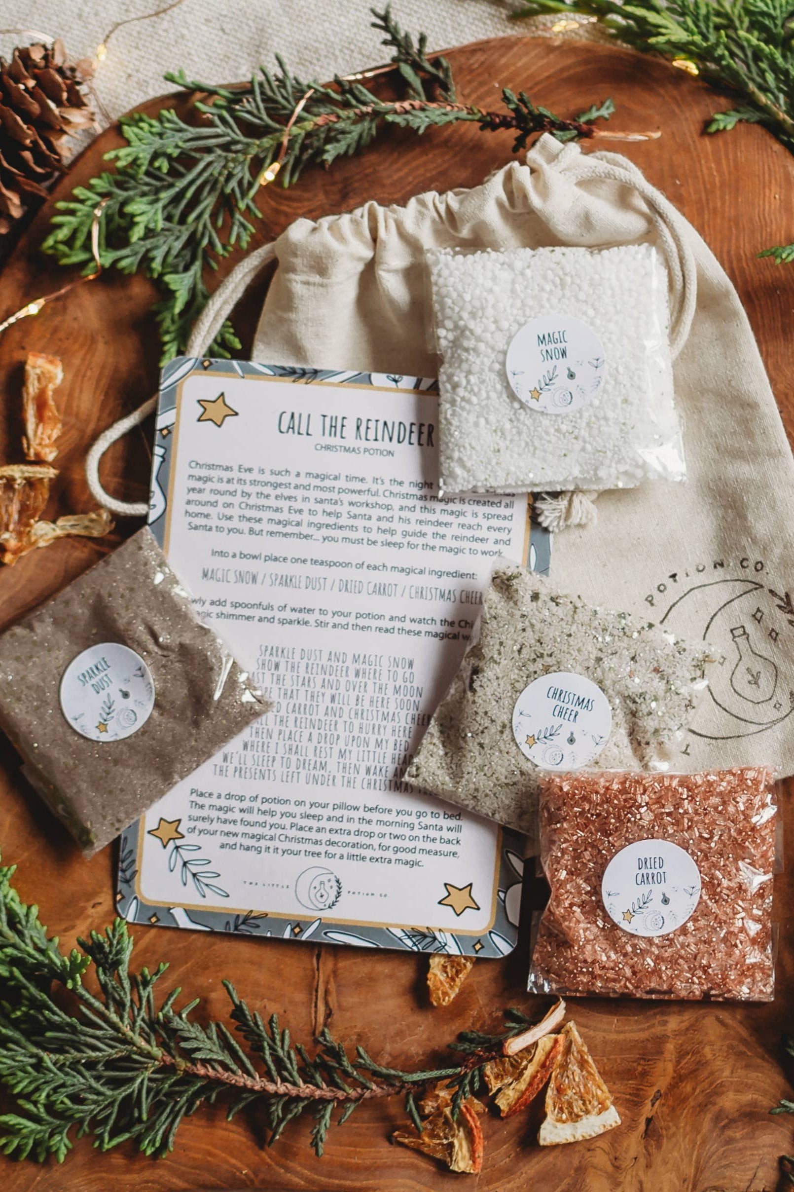 A festive display showcases the Xmas Eve Potion Pouch to Call the Reindeer from THE LITTLE POTION CO, featuring small bags of reindeer-themed magic snow, stardust, and dried carrots on a wooden table. Surrounded by pine branches and orange slices, it includes instructions for maintaining the magical tradition on Christmas Eve.