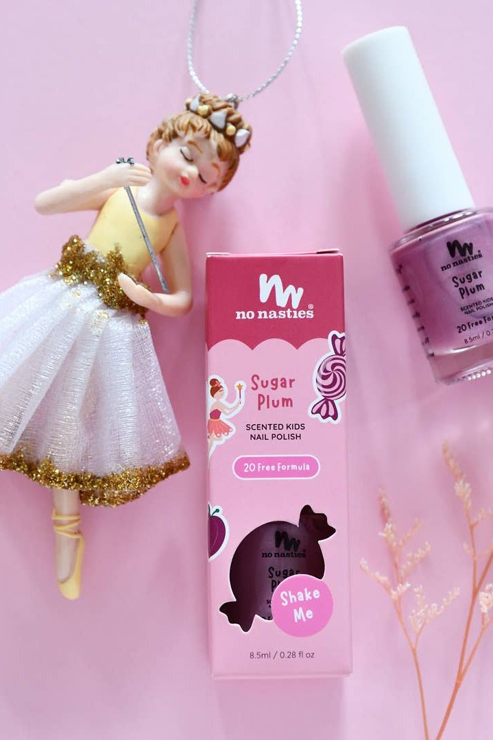 A ballerina figurine in a white and gold dress is placed beside a box of NO NASTIES Sugar Plum Water Based Scented Scratch Off Kids Nail Polish, known for its non-toxic and cruelty-free formula. The corresponding nail polish bottle is positioned nearby against a pink backdrop embellished with tiny dry flowers.