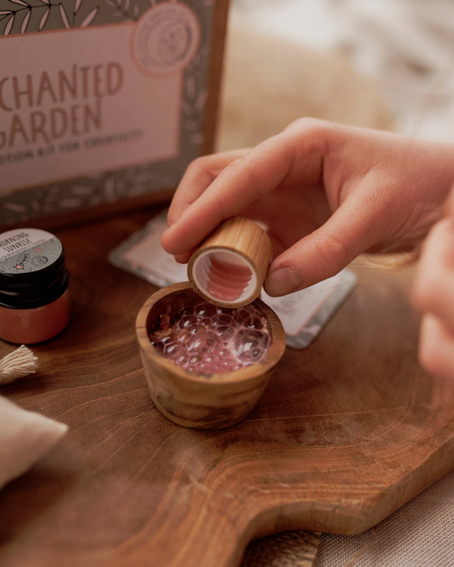 A person pours liquid from a small bottle into a wooden bowl filled with a bubbly mixture. The scene is set on a wooden surface, featuring the "MINI Enchanted Garden Potion Kit (A potion for creativity)" by THE LITTLE POTION CO, complete with mini potion kit jars and packets—perfect for imaginative play.