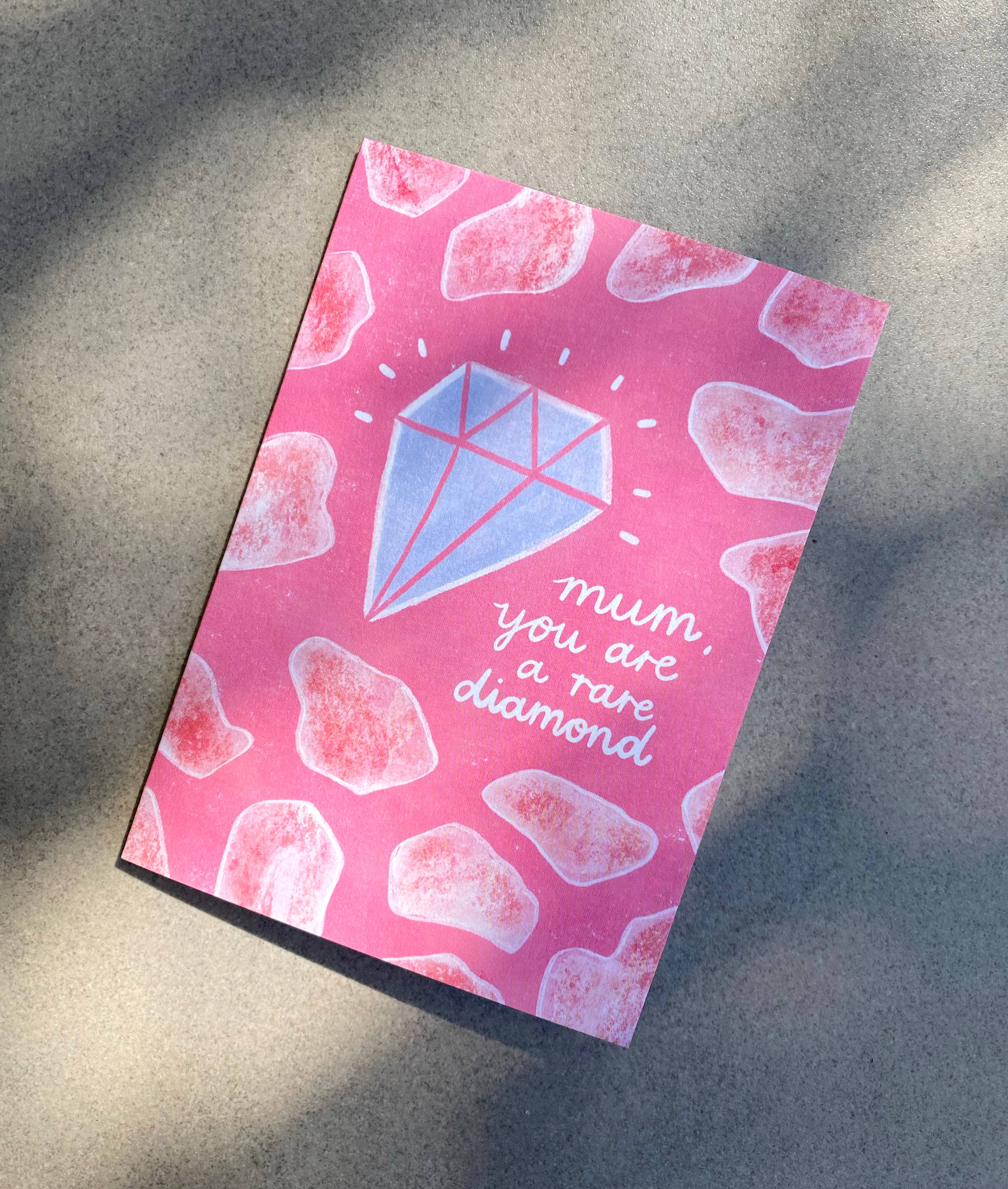 The Mother's Day Greeting Card "Mum, You Are A Rare Diamond" by LAUREN SISSONS STUDIO features a pink background adorned with various pink gemstone shapes. At the center, there is an illustration of a blue diamond accented with white sparkles. The card's message reads, "mum, you are a rare diamond." Shadow patterns appear on the surface where the card is placed. This card is made from recycled paper.