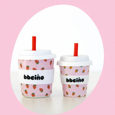 Two BBCINO Reusable BambinoCino Cups 240ml Berry BB come with white lids, silicone straws, and white sleeves adorned with small strawberries. The cups feature a light pink background and are set against a larger pink oval shape. The left cup is double the size of the right one.