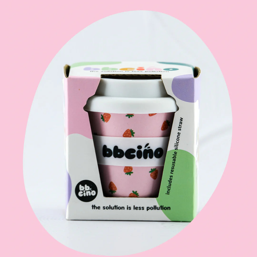 A reusable Babycino Cup 120ml Berry BB from BBCINO, featuring a pink cup with a strawberry pattern made from bamboo fibers, is displayed in a pastel-colored box adorned with white, green, and light purple accents. The box includes a silicone straw and bears the brand name "BBCINO." This environmentally friendly product has a light pink background.