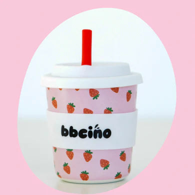 A pink BBCINO Reusable BambinoCino Cup 240ml Berry BB, featuring a white lid and a red silicone straw, beautifully adorned with small strawberry illustrations. It also includes a white sleeve with the text "BambinoCino reusable cup" in black letters. This cup, twice the size of your standard option, is centered against a light pink background.