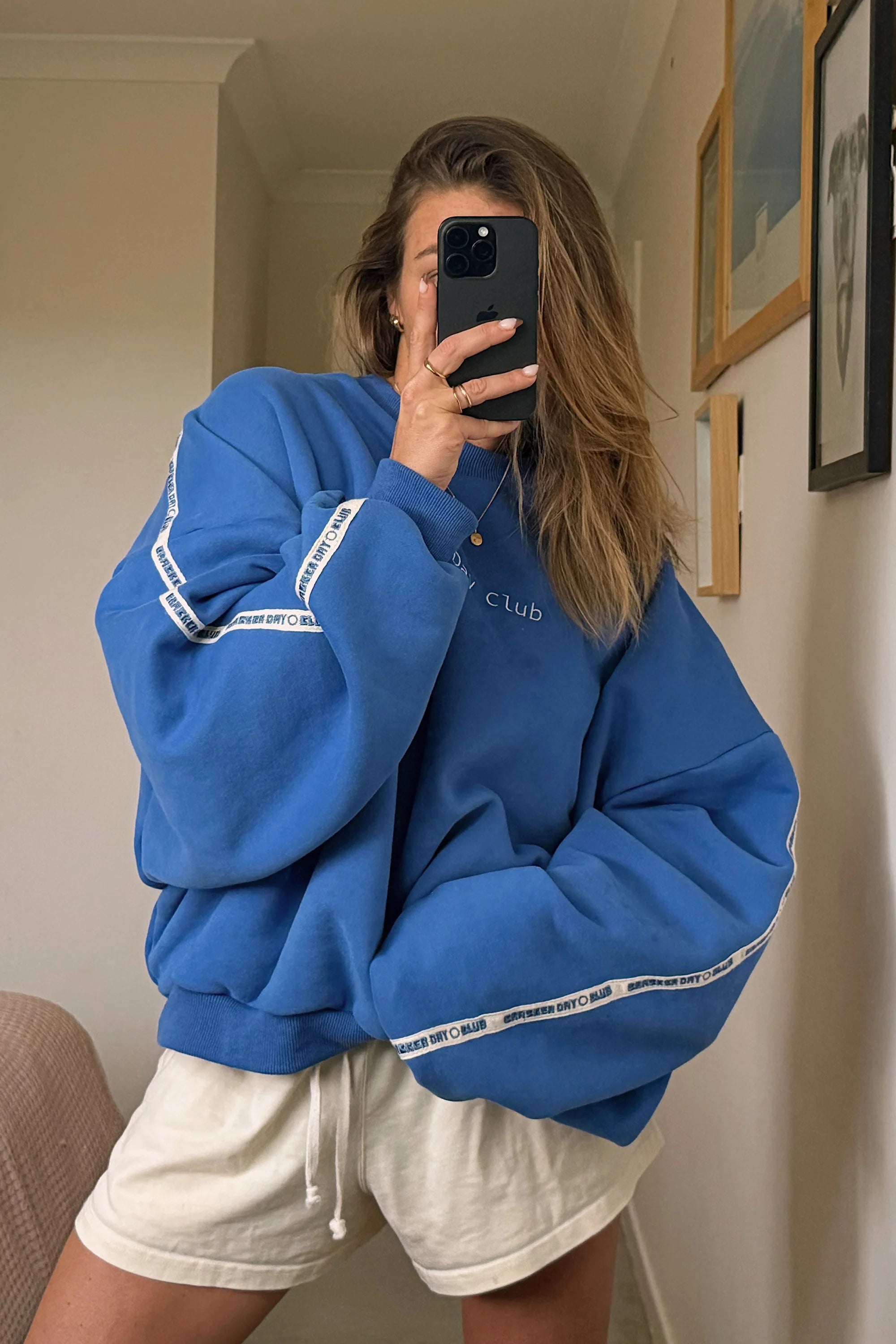 Wearing the Cracker Day Club - Cloud Racer Sweater Blue by CRACKER DAY CLUB with white shorts, a person snaps a mirror selfie, partially hiding their face with a phone. Framed pictures hang on the light-colored wall behind them.
