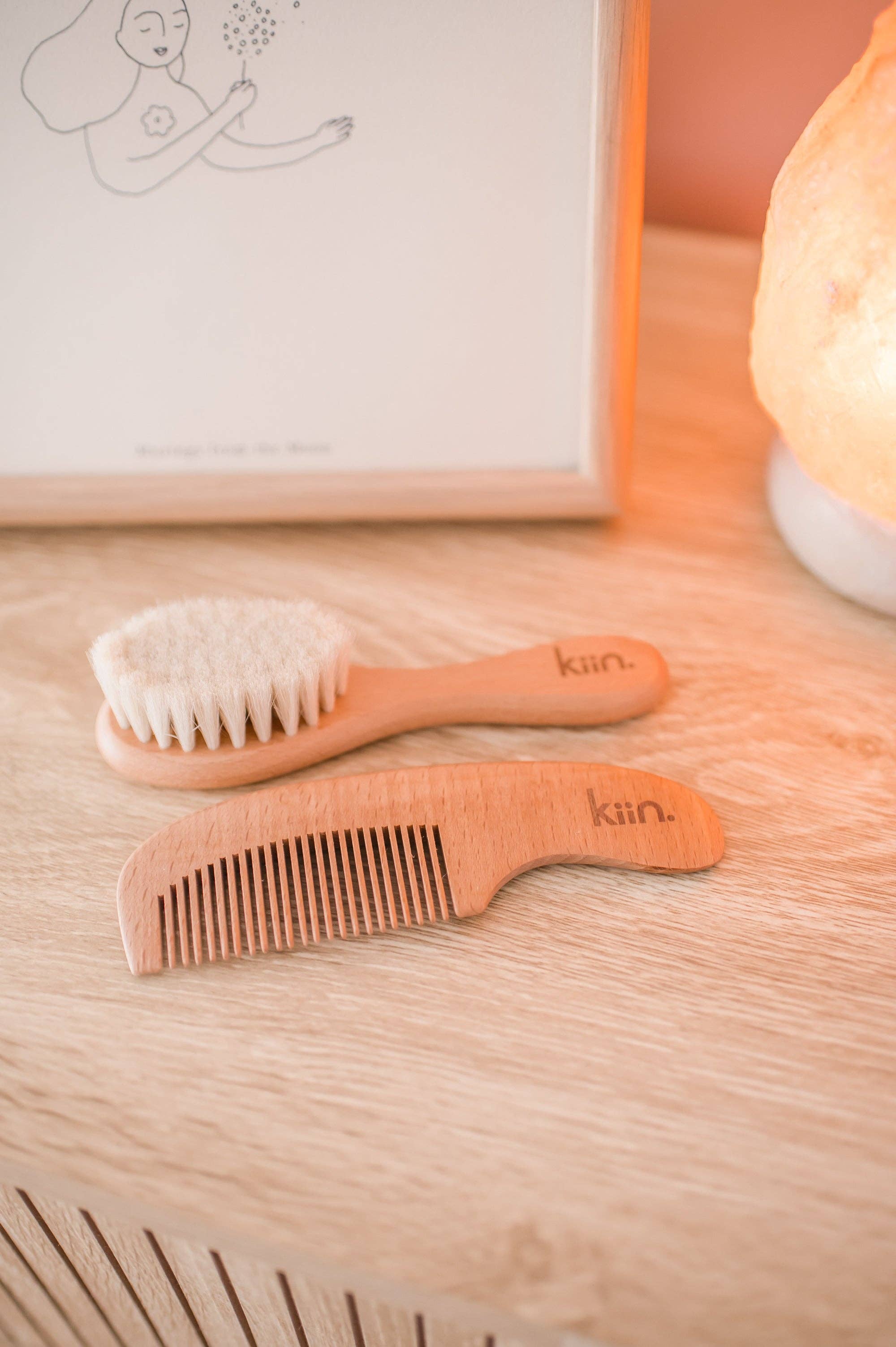 A Wooden Baby Brush and Comb Set from KIIN BABY, featuring gentle goat wool, sits on a wooden surface. Behind it are a softly glowing lamp and a minimalist framed illustration, creating an eco-friendly ambiance.