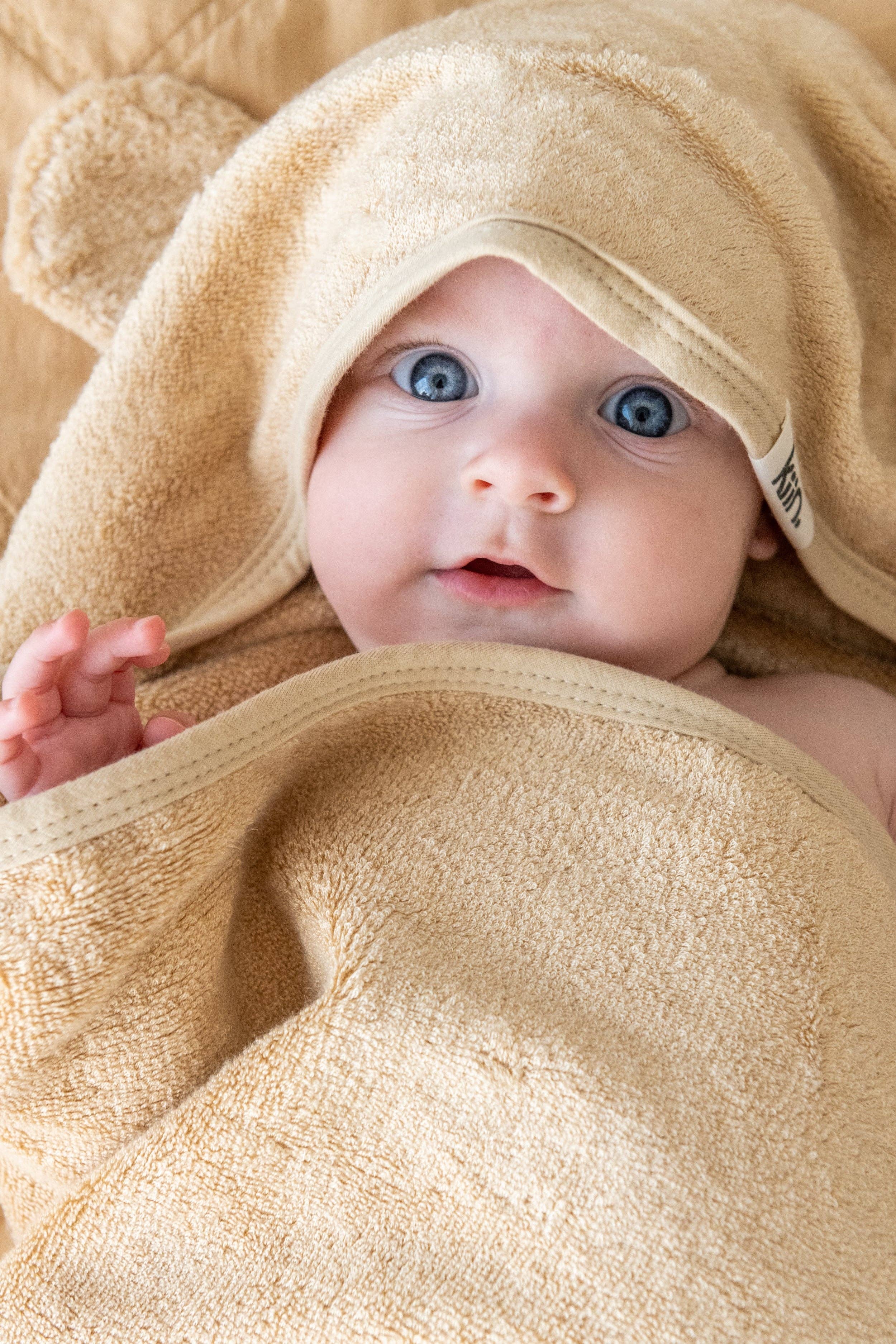 A baby with bright blue eyes is wrapped in a KIIN BABY Hooded Towel Ivory featuring bear ears. The hypo-allergenic bamboo fabric gently covers the baby's head and body, creating an endearing scene.