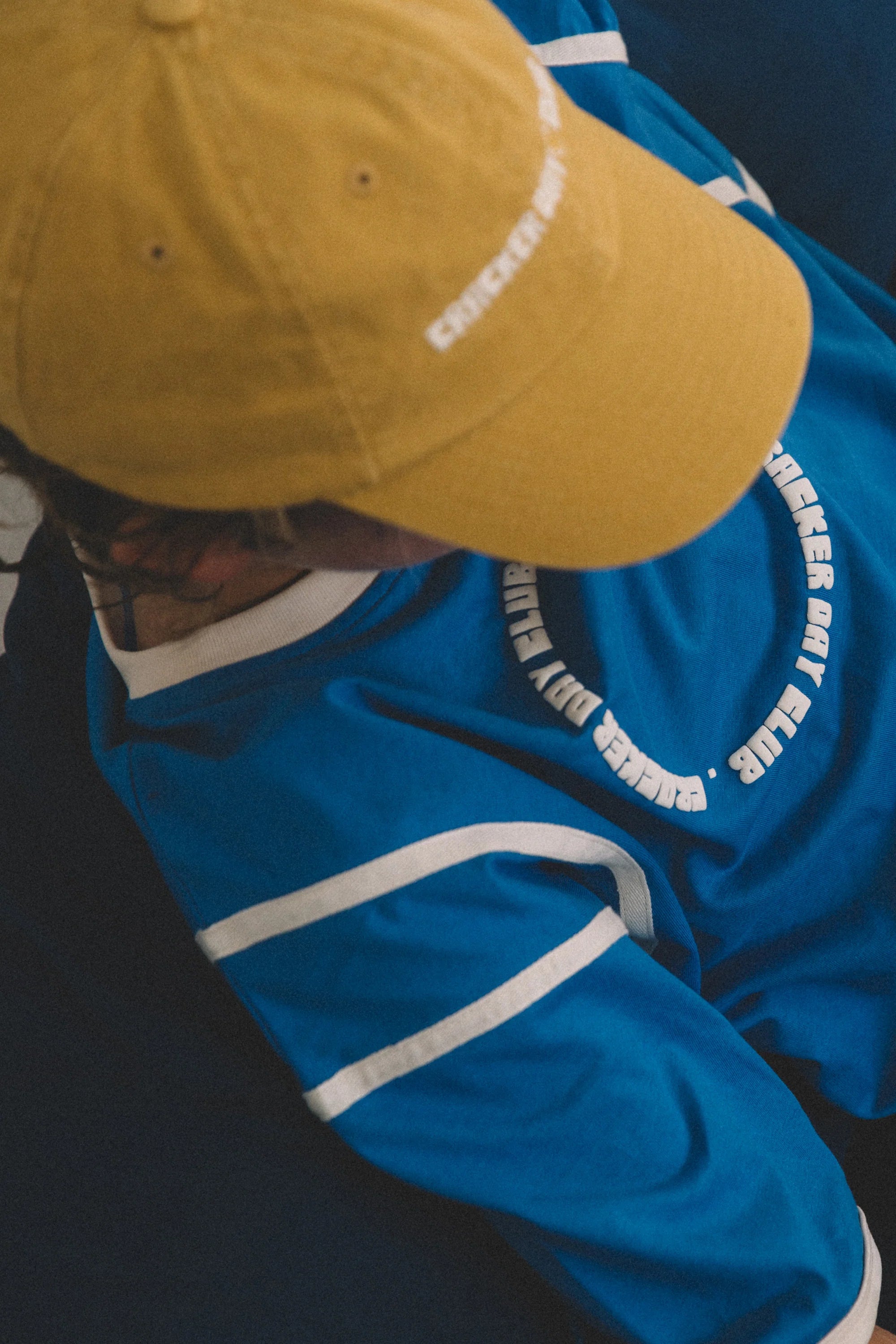 A child wears a yellow cap and a CRACKER DAY CLUB Cracker Day Club - Kids Racer Club T-Shirt Blue with white stripes and curved white text on the back, seen from above against a dark, blurred background.