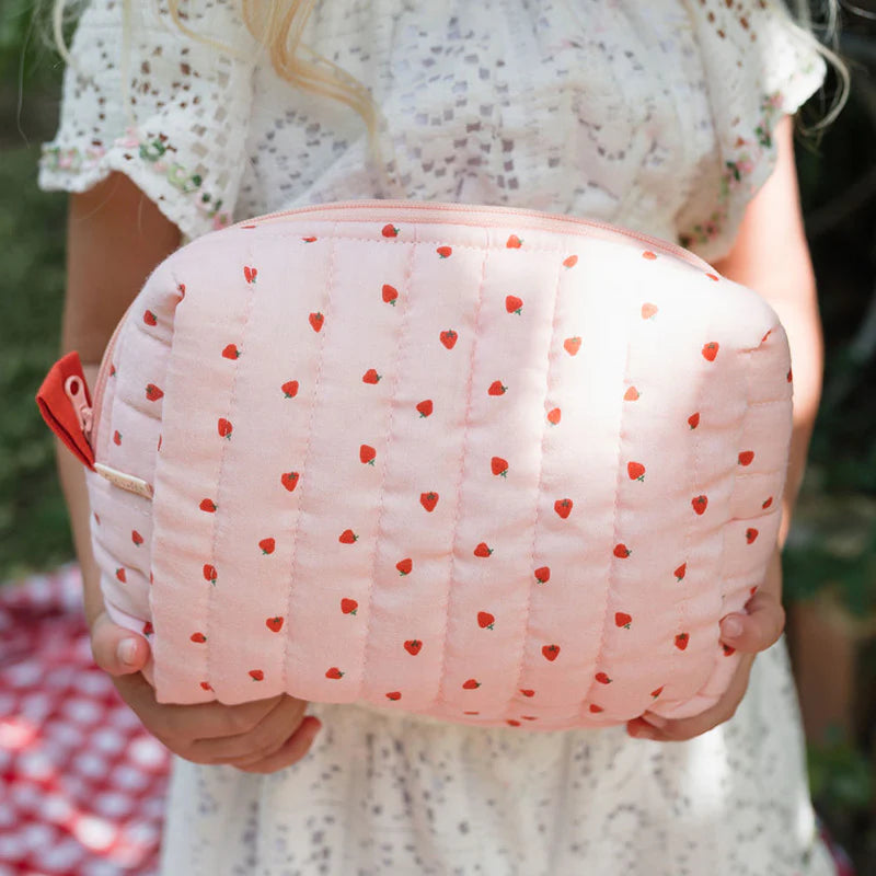A person in a white eyelet dress holds the Olli Ella - Quilted Toiletries Bag Strawberry by OLLI ELLA, surrounded by greenery while sitting on a red-checkered blanket.