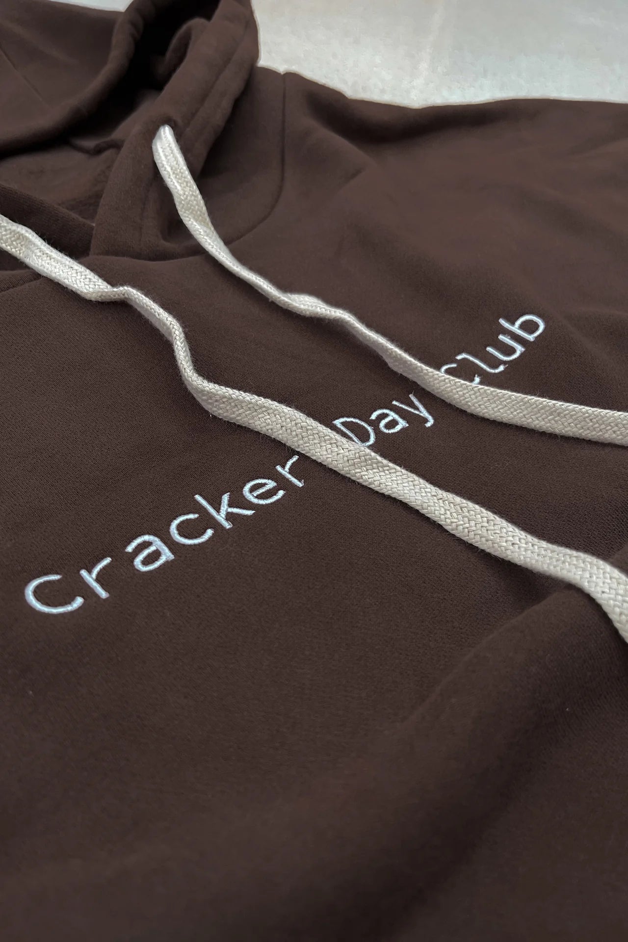 A close-up of the CRACKER DAY CLUB Cloud Racer Hoodie Chocolate, featuring an oversized fit, white drawstrings, and "Cracker Day Club" embroidered in white on the chest. Made from heavyweight terry cotton for comfort and style.
