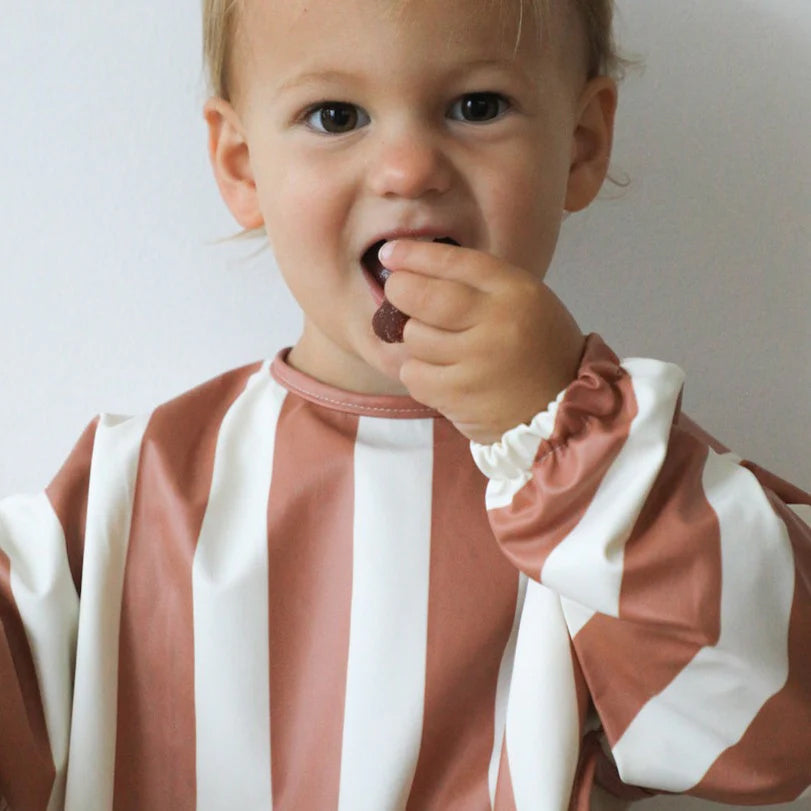 A young boy wearing the ROMMER Smock Bib in Cinnamon Stripe eating a lolly.