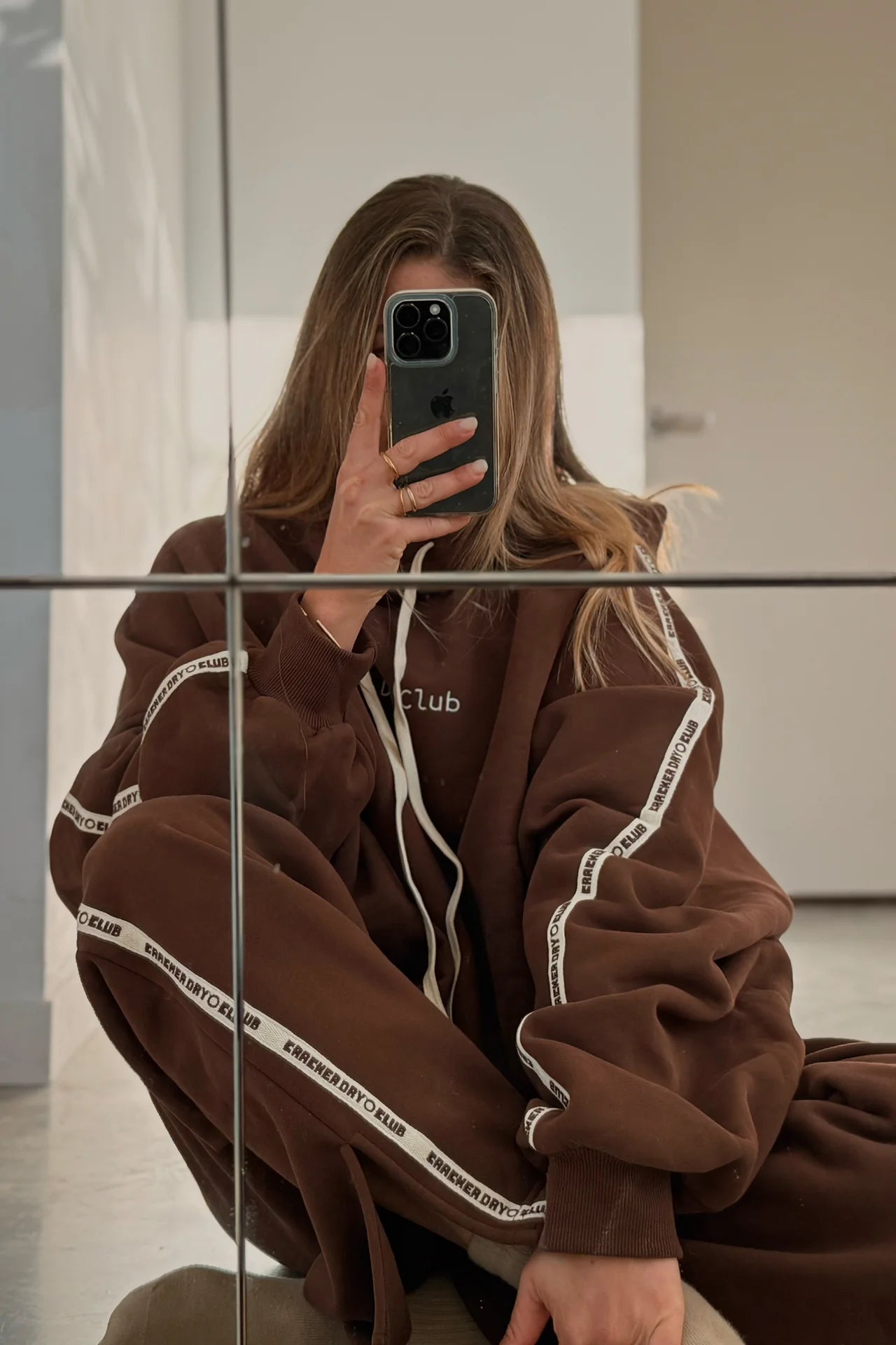 A person in a CRACKER DAY CLUB Cloud Racer Hoodie Chocolate takes a mirror selfie on the floor. The hoodie features white stripes, their face is partially hidden by the phone, and their long hair falls loosely around them.