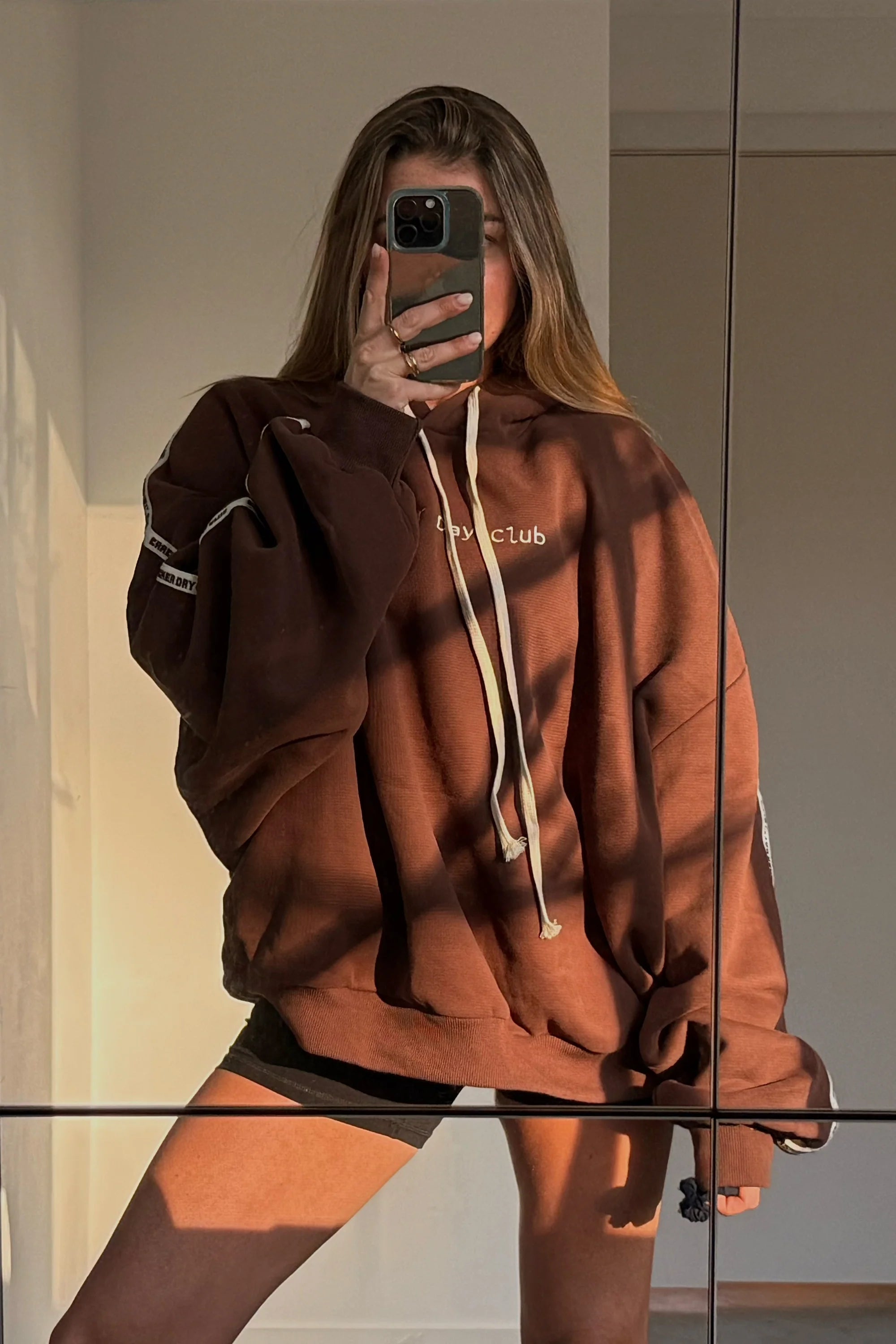 A person with long hair takes a mirror selfie in the CRACKER DAY CLUB Cloud Racer Hoodie Chocolate, featuring heavyweight terry cotton, white drawstrings, and black shorts. Sunlight and shadows fall across them and the wall.