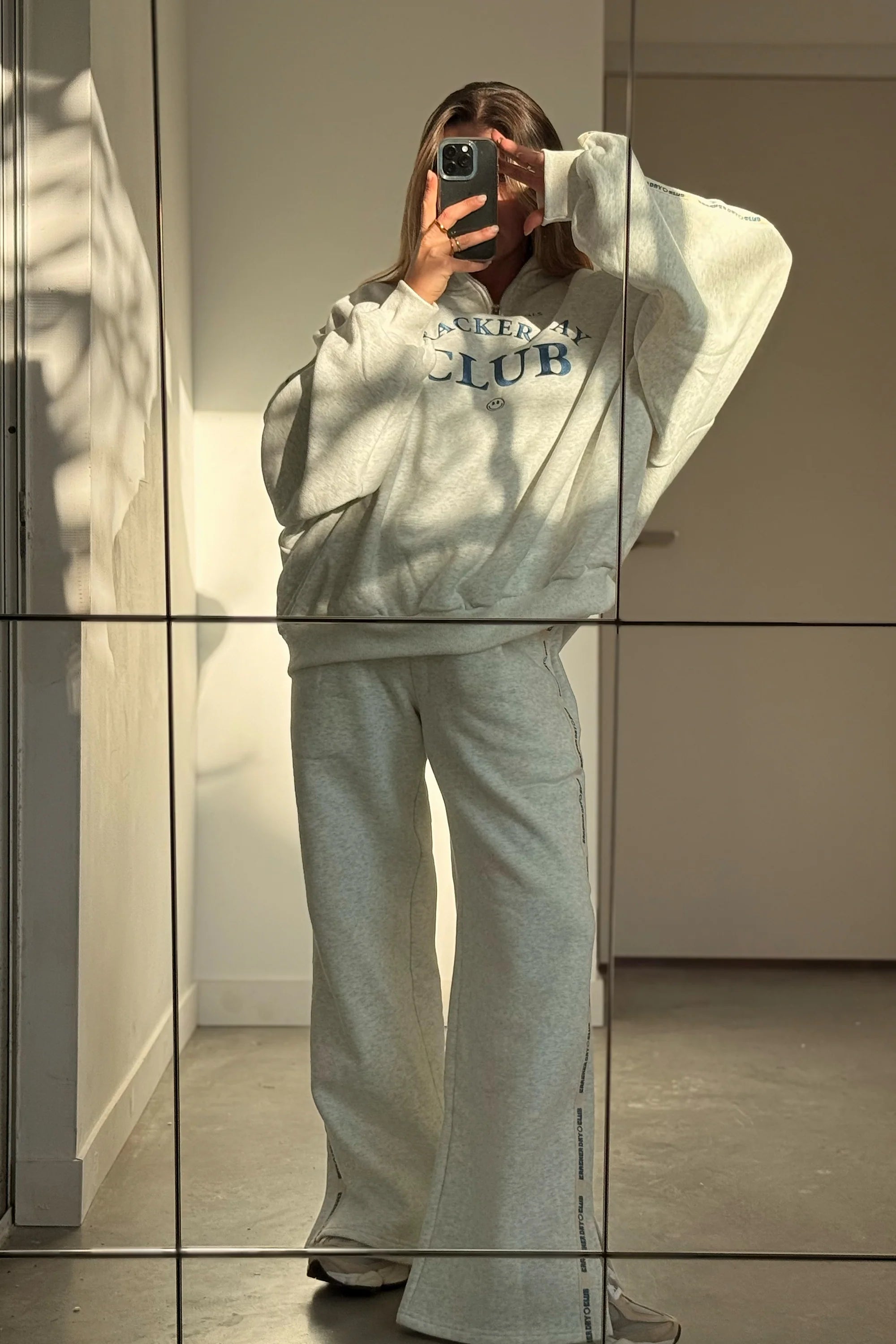 Wearing CRACKER DAY CLUB’s Cloud Racer Sweatpants in Grey Marle, a person snaps a mirror selfie in a sunlit room, soft shadows and light playing across the wall behind them.
