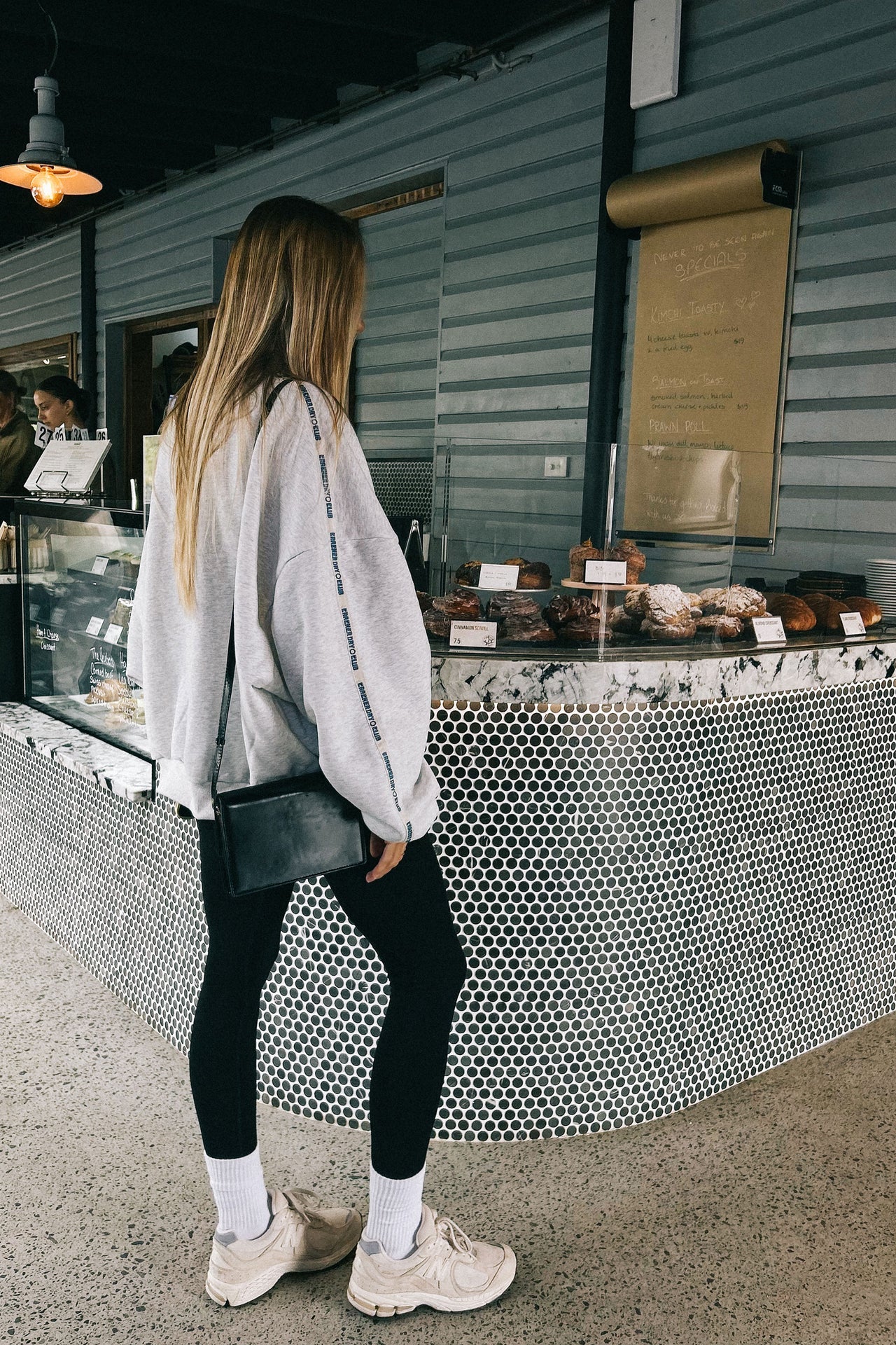 A woman with long hair, wearing a CRACKER DAY CLUB Cloud Quarter Zip Sweater in Grey Marle, black leggings, white socks, and sneakers, stands by a pastry-filled bakery counter in a modern café.