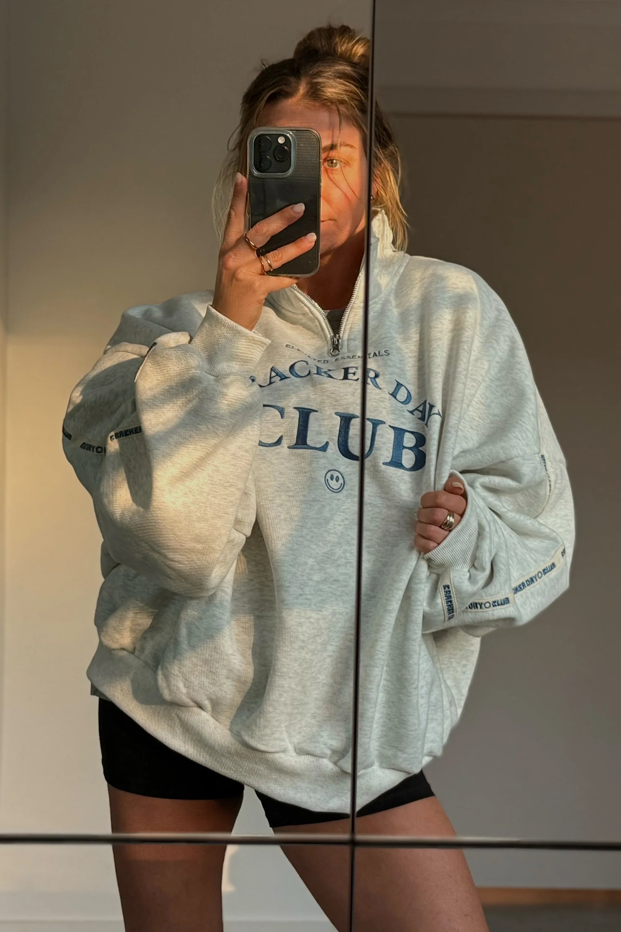 Wearing the CRACKER DAY CLUB Cloud Quarter Zip Sweater in Grey Marle, someone takes a mirror selfie, partially hiding their face with their phone. The heavyweight cotton’s oversized fit catches sunlight and indoor shadows.