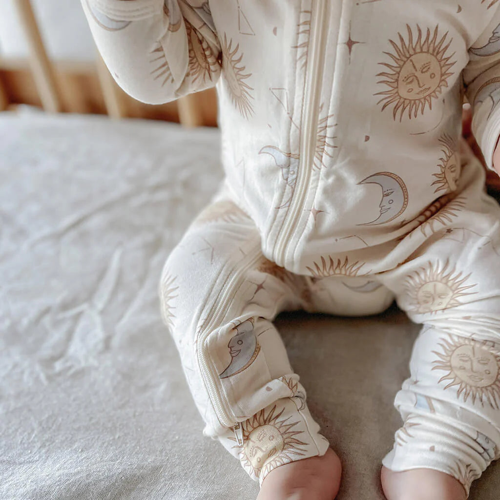 A baby in a cream-colored Celestial Zip Jumpsuit from BAM LOVES BOO, made with a blend of bamboo and organic cotton, is adorned with a celestial pattern of suns and moons while seated on a soft surface. The arms and torso are visible, with the legs crossed at the ankles, framed by a wooden crib railing.