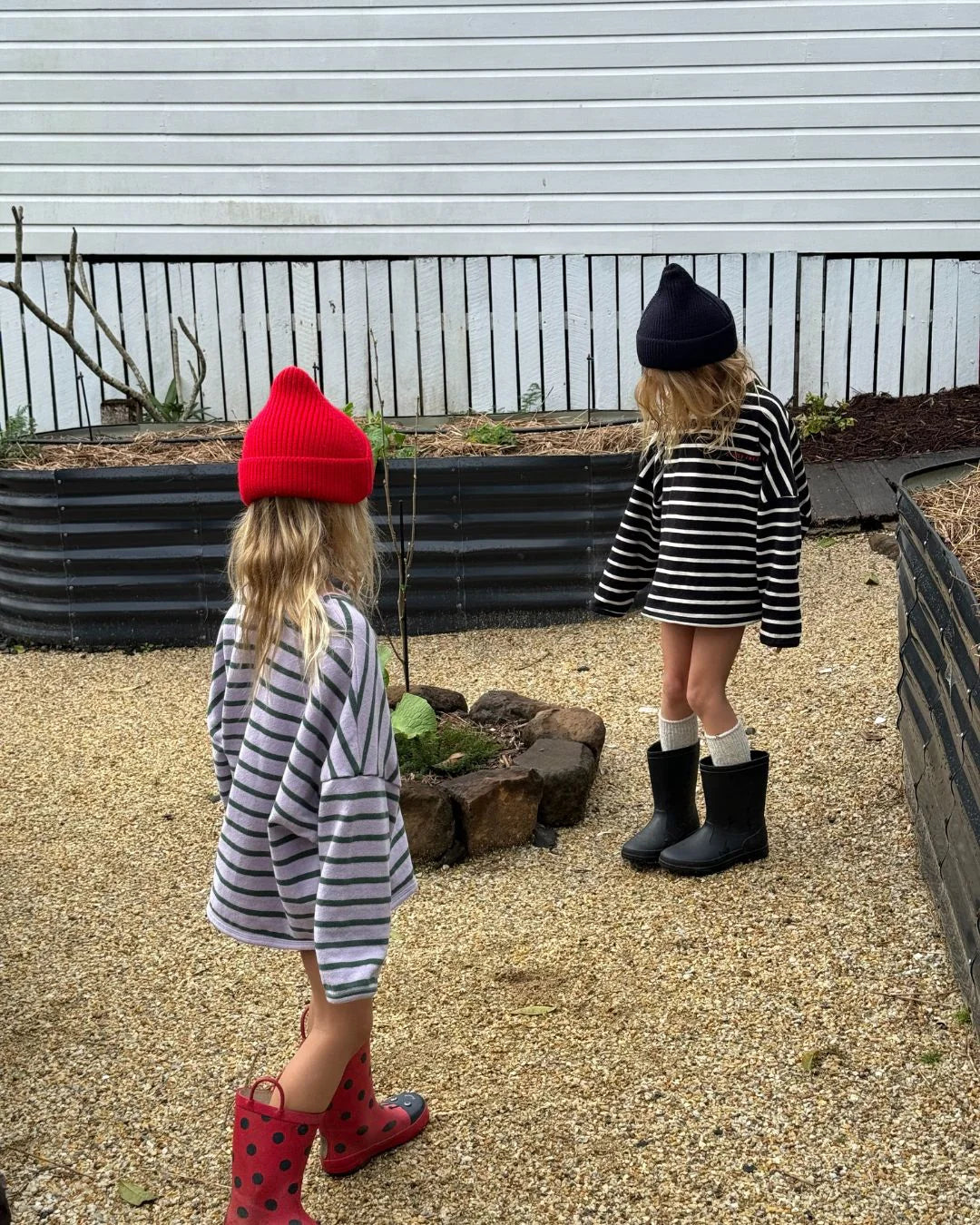 Two children stand outside by a small rock circle, wearing oversized ZIGGY ZAZA Row Row Pullover Lilac Stripe shirts with beanies and boots—one in red boots and hat, the other in black boots and hat.