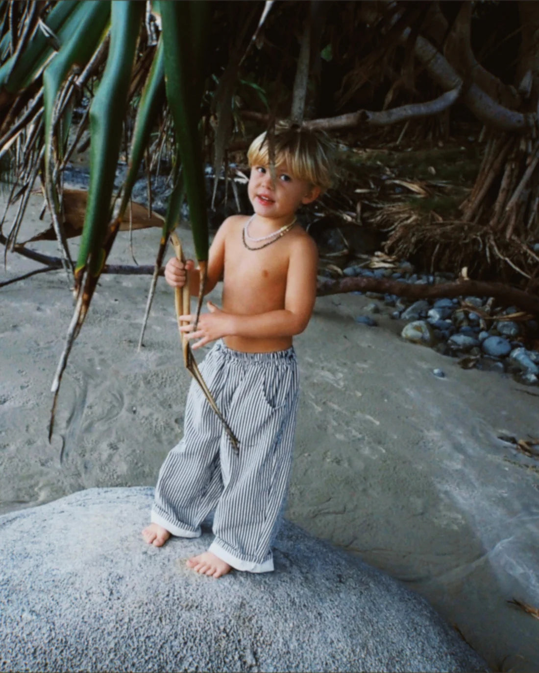 A young child with blonde hair, barefoot on a beach rock, wears ZIGGY ZAZA’s Ziggy Zaza - Casper Pant Navy Stripe and a beaded necklace, holding a leafy branch with sand and trees in the background.