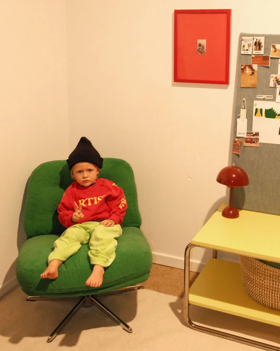 A young child in a relaxed fit, 100% cotton fleece Ziggy Zaza - Artist Pullover Racer Red by ZIGGY ZAZA and green pants sits barefoot on a green chair beside a yellow table with a red lamp and nearby bulletin board.