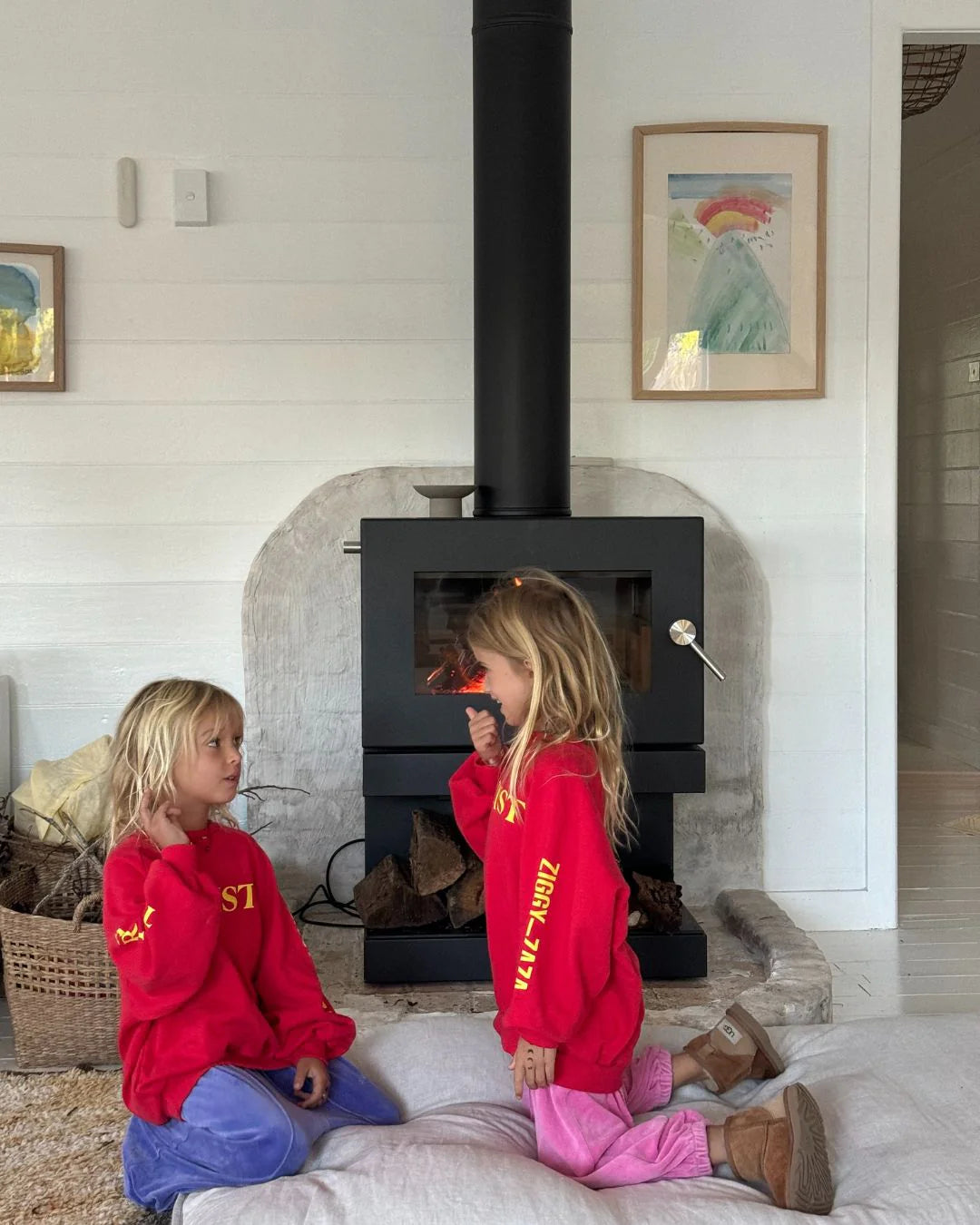 Two young children in matching ZIGGY ZAZA Ziggy Zaza - Artist Pullover Racer Red toast marshmallows by a black wood-burning stove, cozy in a white-walled room. The relaxed fit and 100% cotton fleece keep them comfy and warm.