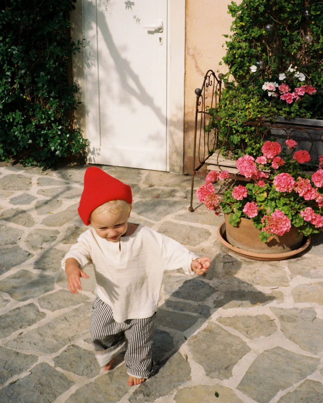A small child in a red beanie, white shirt, and ZIGGY ZAZA Ziggy Zaza - Casper Pant Navy Stripe walks barefoot on a stone patio by pink flowers, with greenery and a white door in the background.