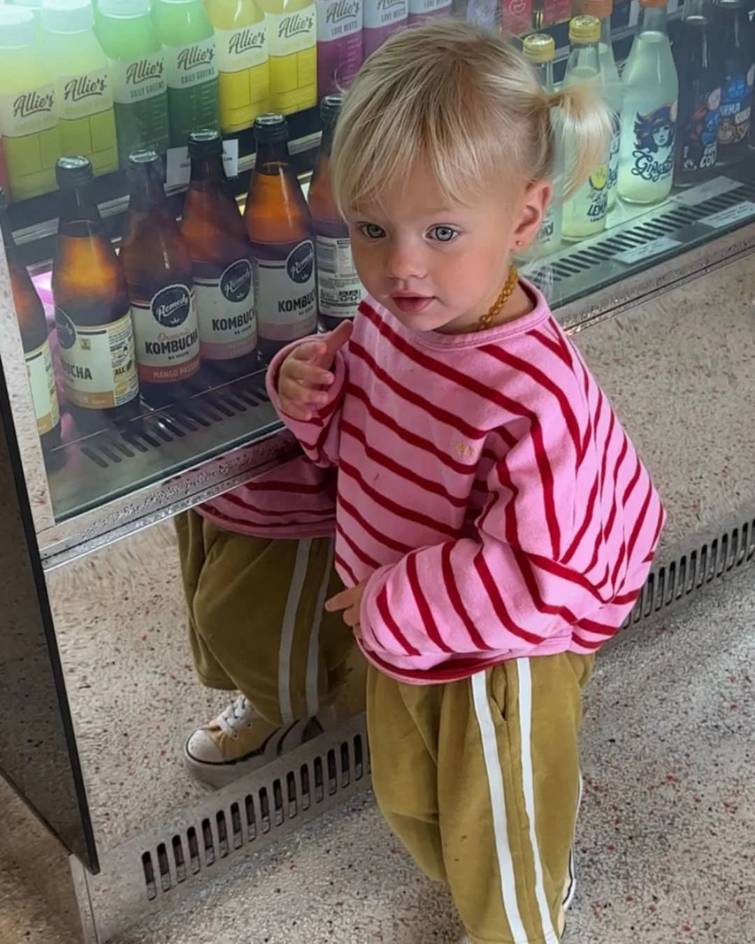 A young child with blonde pigtails wears the ZIGGY ZAZA Row Row Pullover Rose Stripe and loose brown pants, standing in front of a refrigerated display filled with bottles of drinks.