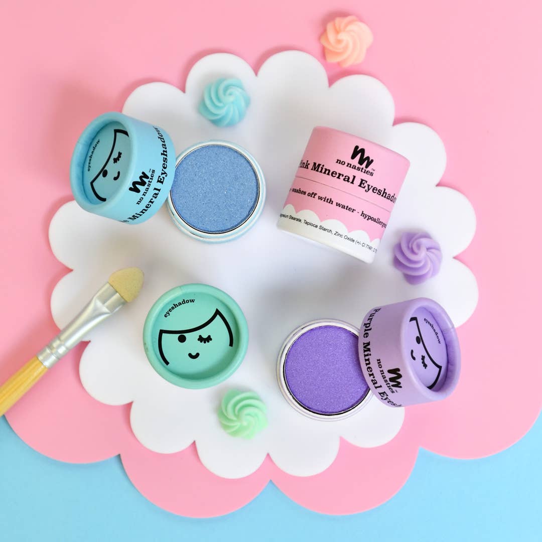 A delightful arrangement of Kids Eco Natural Pressed Eyeshadow & Blush by no nasties kids, featuring vibrant pastel hues, sits on a pink and blue backdrop with a charming white scalloped border. Each container showcases an adorable face illustration, made from natural ingredients and certified cruelty-free. A small brush rests on the left side, surrounded by cute candy-shaped decorations to complete the playful scene.