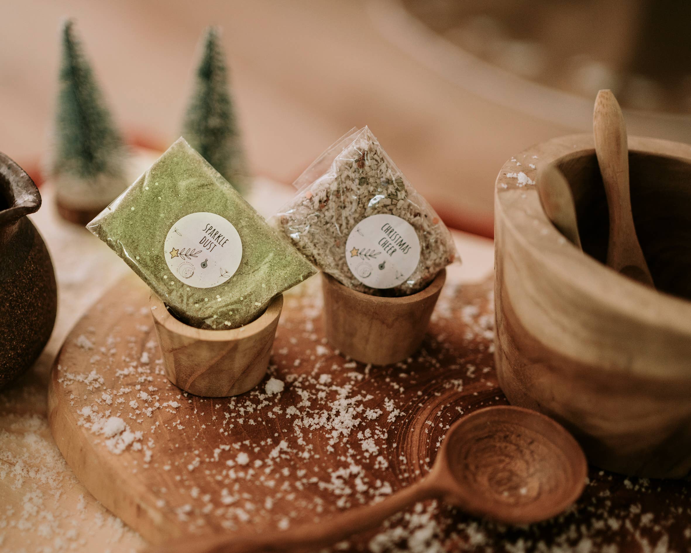 Two small packages, labeled as "Xmas Eve Potion Pouch to Call the Reindeer," from THE LITTLE POTION CO, rest on wooden stands atop a wooden board, reminiscent of a Christmas potion kit. A wooden mortar with a pestle and spoon are nearby. Snow-like decorations and mini evergreen trees add a festive touch, evoking the charm of a magical tradition.