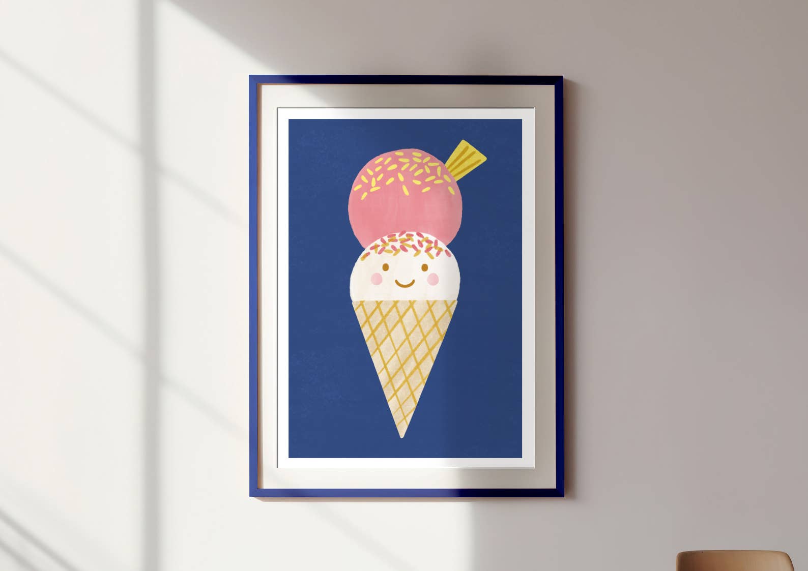 The Ice Cream Wall Art Print by LAUREN SISSONS STUDIO is a framed contemporary piece showcasing a minimalist and adorable illustration of an ice cream cone with two scoops—one pink with sprinkles and one white. The smiling ice cream stands out against a dark blue background. Printed on high-quality art paper, this picture is displayed on a light-colored wall.