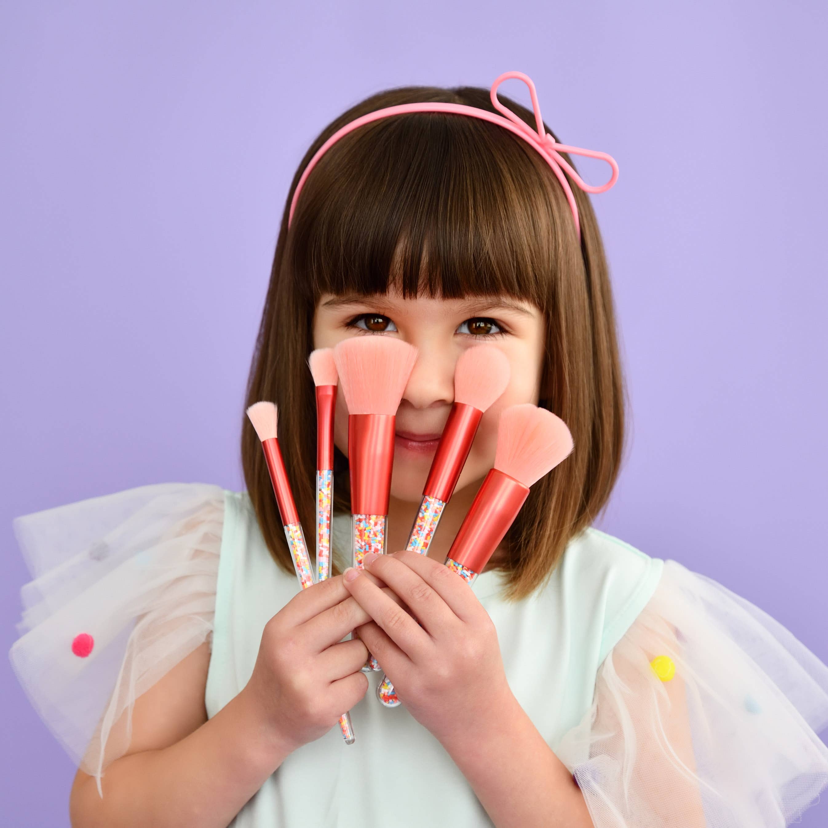 A young girl with straight brown hair, wearing a light blue dress adorned with colorful pom-poms and a pink bow headband, holds the "Twinkle Sprinkle Brush Set" from "no nasties kids" with its pink bristles and decorative handles in front of her face, set against a purple background.