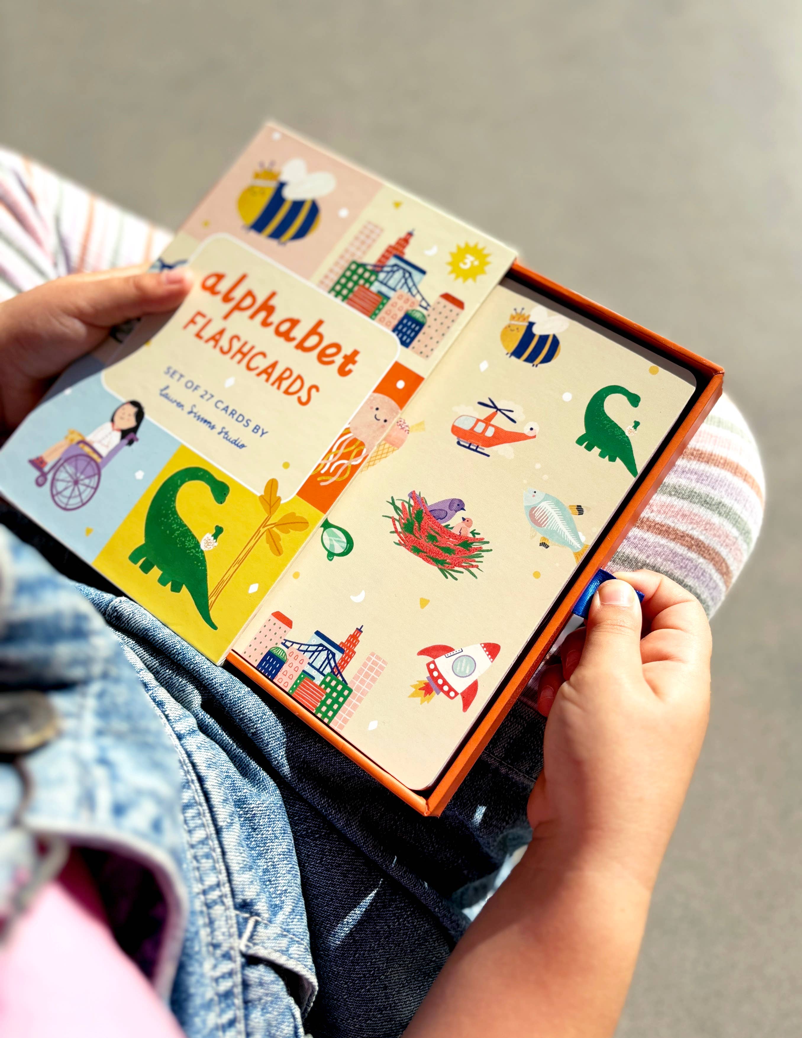 A child's hands holding a colorful box of A-Z Illustrated Alphabet Flashcards from LAUREN SISSONS STUDIO. The box features playful illustrations of various items such as a dinosaur, a bird's nest, a hot air balloon, and buildings. The child is sitting, wearing a striped outfit and denim jacket. These educational toys for children are laminated for durability.