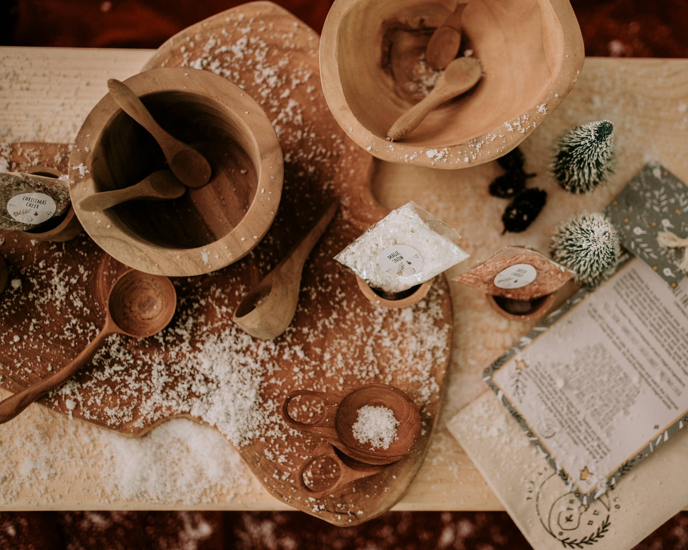 A rustic table setting featuring wooden bowls and spoons, with artificial snow sprinkled across the surface, multiple sachets of spices, and two small decorative Christmas trees. An open book with festive illustrations sits next to THE LITTLE POTION CO's Xmas Eve Potion Pouch to Call the Reindeer, perfectly capturing the essence of a magical tradition.